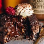 A baked chocolate pudding (lava cake) is open revealing the gooey chocolate centre, it has cream and a raspberry on top with small pieces of grated chocolate.
