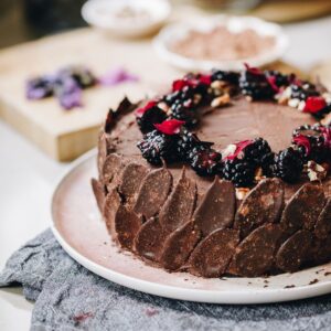 A whole vegan chocolate cake is plated on a pink plate. It is decorated in dark chocolate petals and topped with berries.