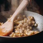 In a silver pan is finely diced pieces of pear that are steaming with spices on it, there is steam coming out of the pan along with a wooden spoon.