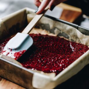 Bright chia raspberry and lemon jam is being spreading in to a slice tin with a clue spatula.