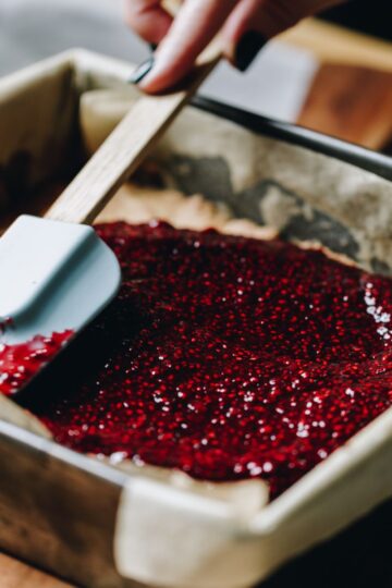 Bright chia raspberry and lemon jam is being spreading in to a slice tin with a clue spatula.