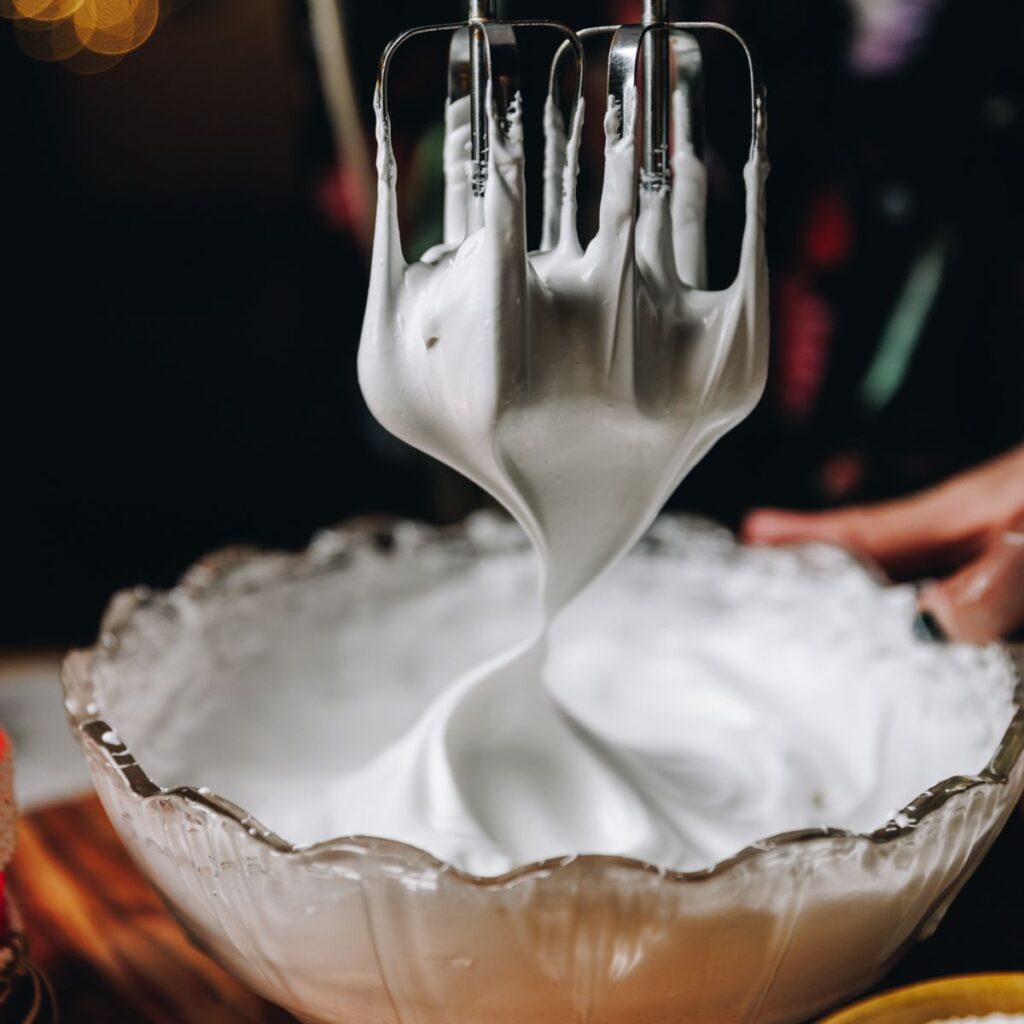 In a glass bowl is Swiss meringue, hand beater whisk attachments are coming out of the meringue revealing stiff peaks.