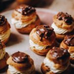 Spiced pear choux buns have been filled with piped whipped cream and topped with a touch of chocolate, they are sitting on a pink plate.