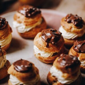 Spiced pear choux buns have been filled with piped whipped cream and topped with a touch of chocolate, they are sitting on a pink plate.