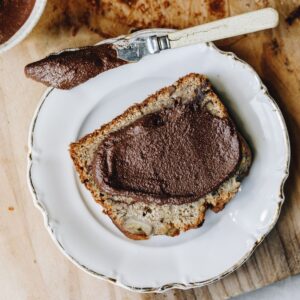 Chocolate almond butter is spread on to a piece of spiced banana loaf, the knife also has heaps of the butter on it. It is all sitting on a white plate, on a wooden board.