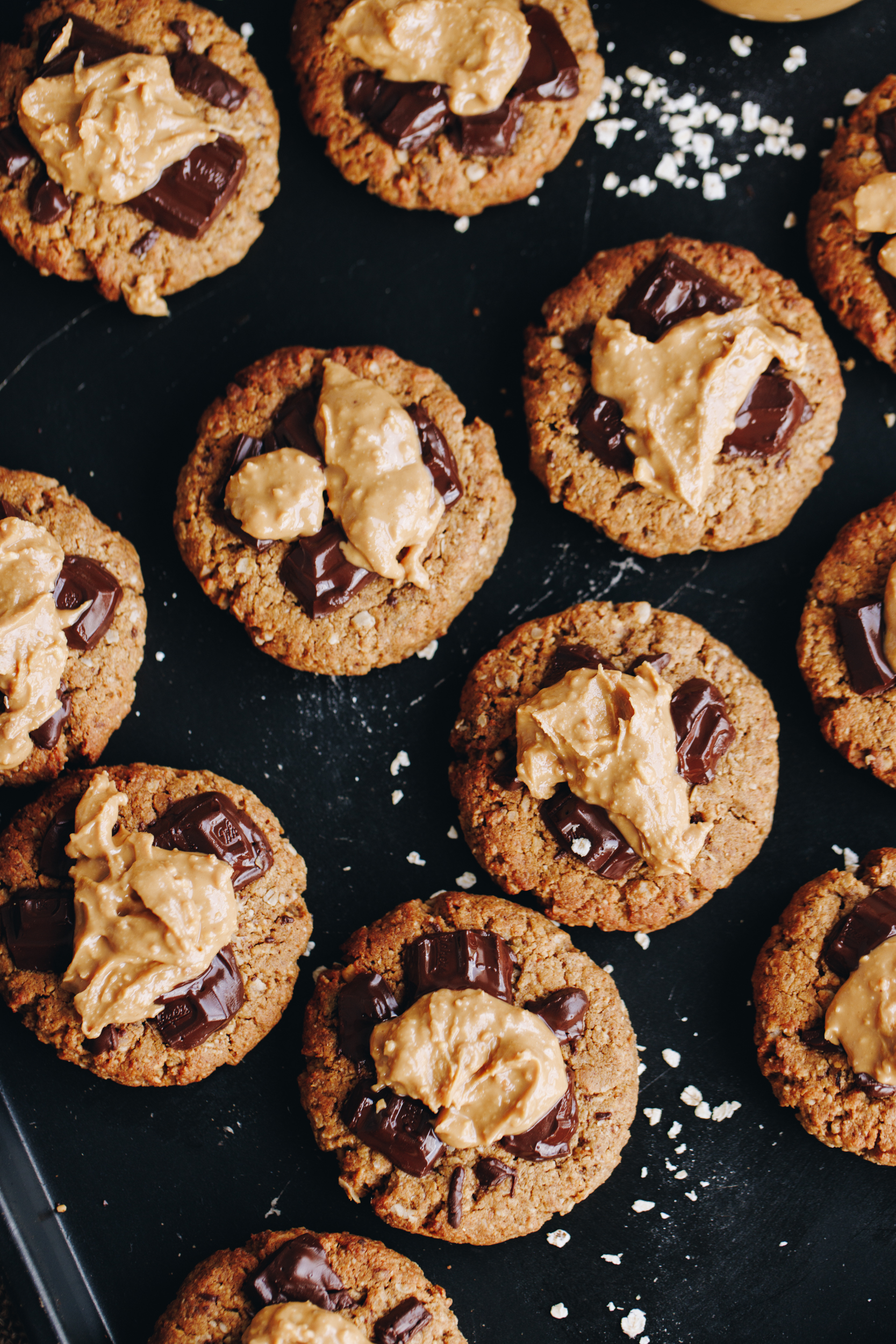 On a black tray are freshly baked wheat-free PB breakfast cookies. Th stop shot shows the cookies with chocolate on the top and peanut butter spread in the centre, there is a scattering of flaky salt on the tray. 