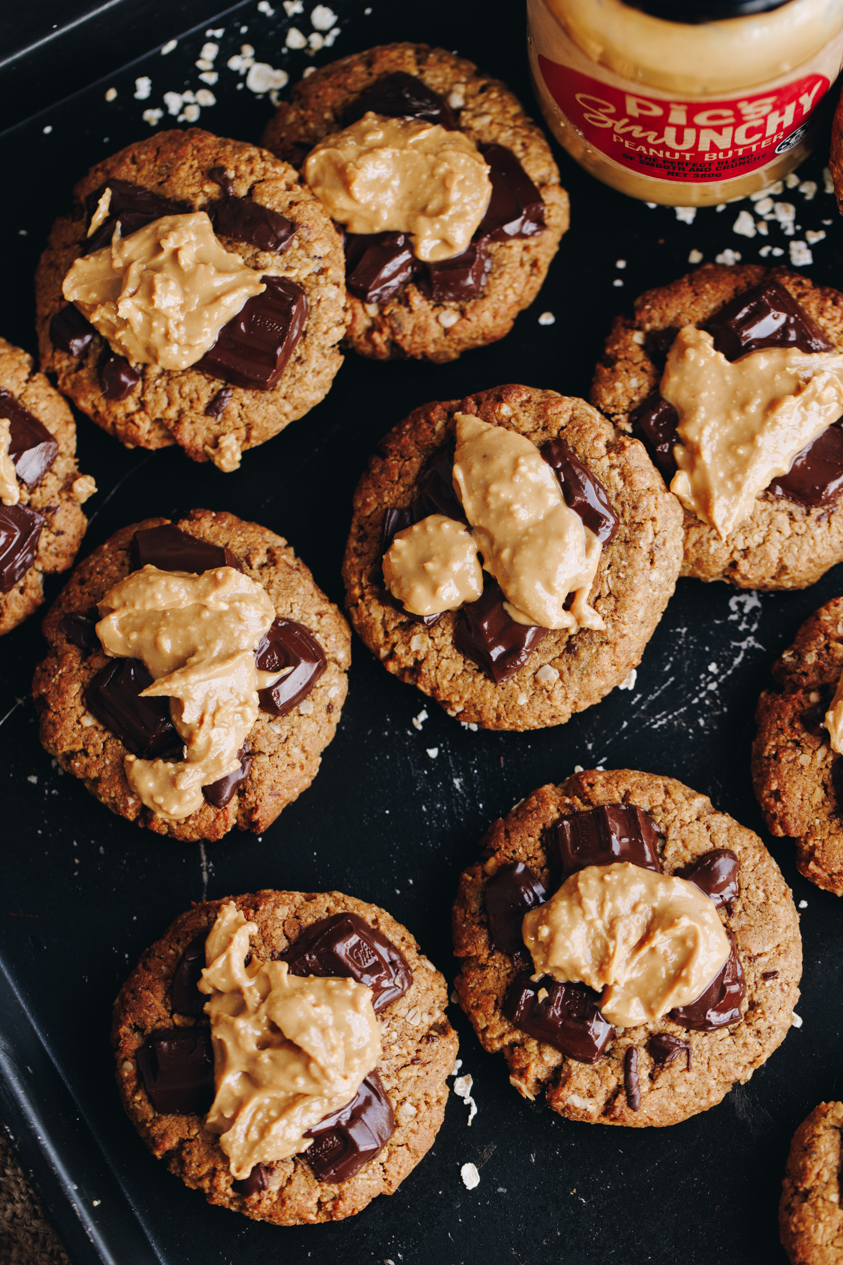 On a black tray are freshly baked wheat-free PB breakfast cookies. Th stop shot shows the cookies with chocolate on the top and peanut butter spread in the centre, there is a scattering of flaky salt on the tray. A Pic's Smunchy peanut butter jar in the corner.  