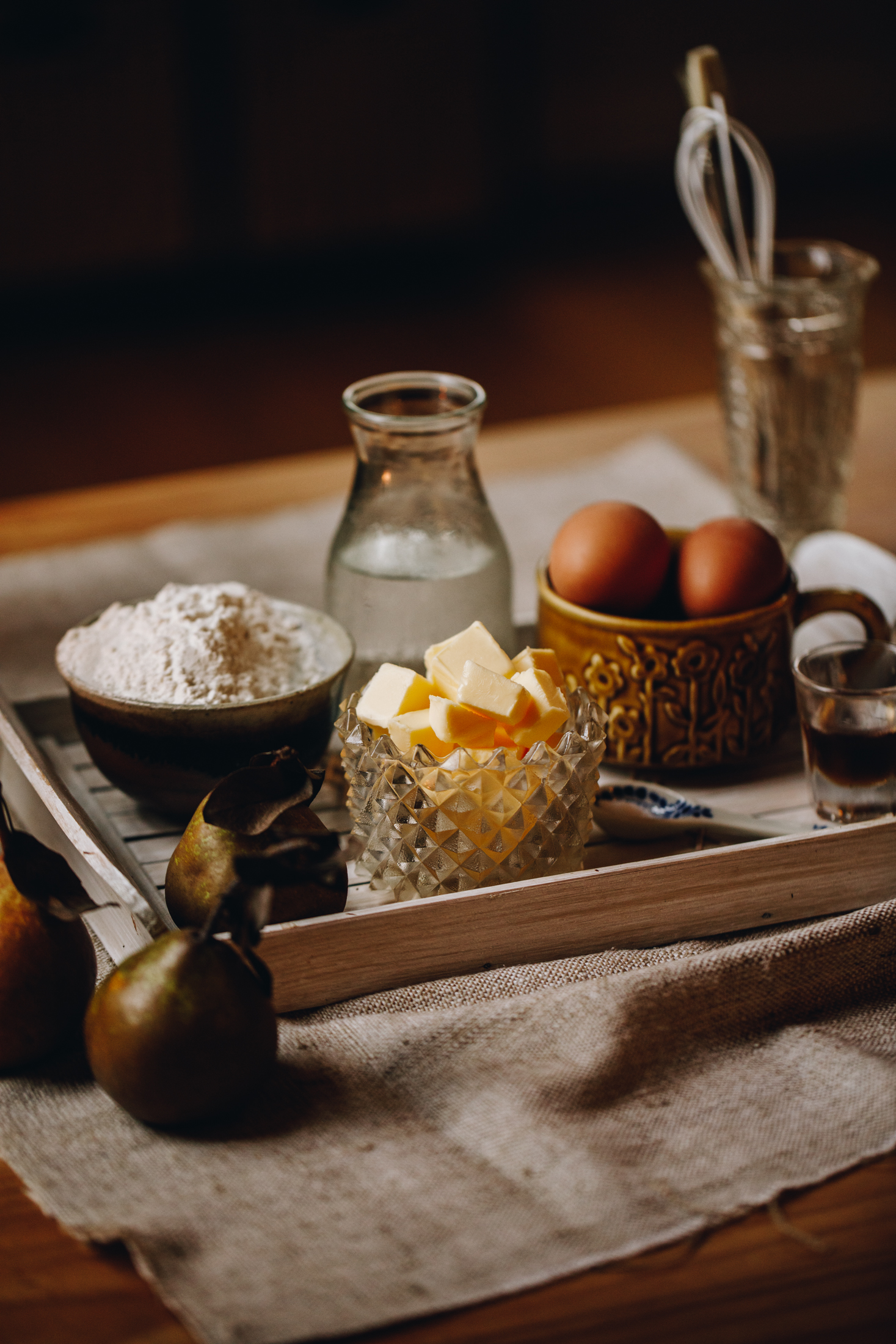 A white wooden tray sits on a beige cloth on a wooden table. Next to the white tray is two pears with stalks and leaves. On the tray is flour, butter, eggs, vanilla and water in vintage cups and glasses. A glass vase with a small whisk is in the background.