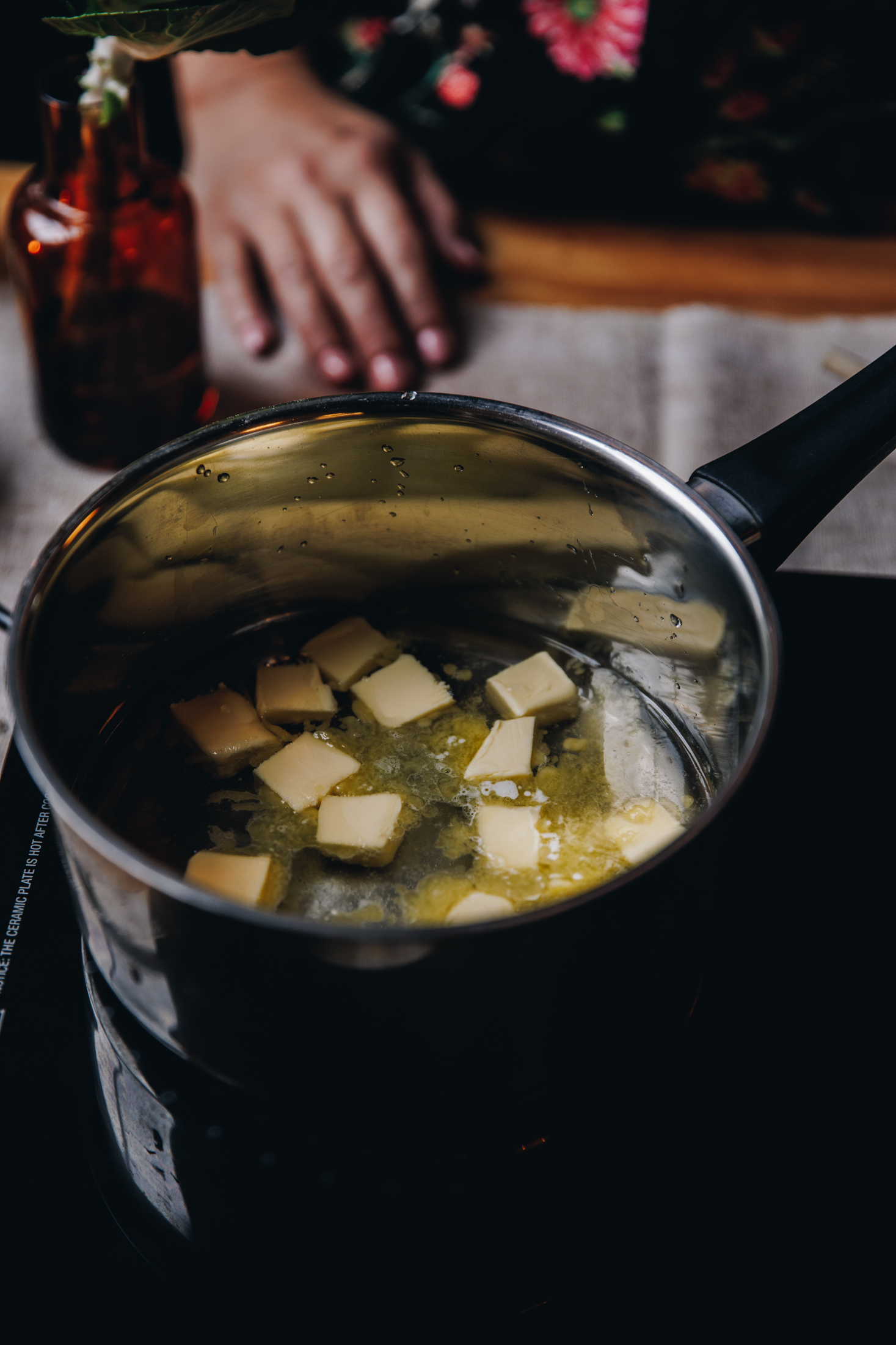A black portable stove sits on a grey cloth on a wooden table. On the stove is a silver pot with a black handle. In the pot is water and butter melting in it. Naomi's hand is resting on the table behind it. 