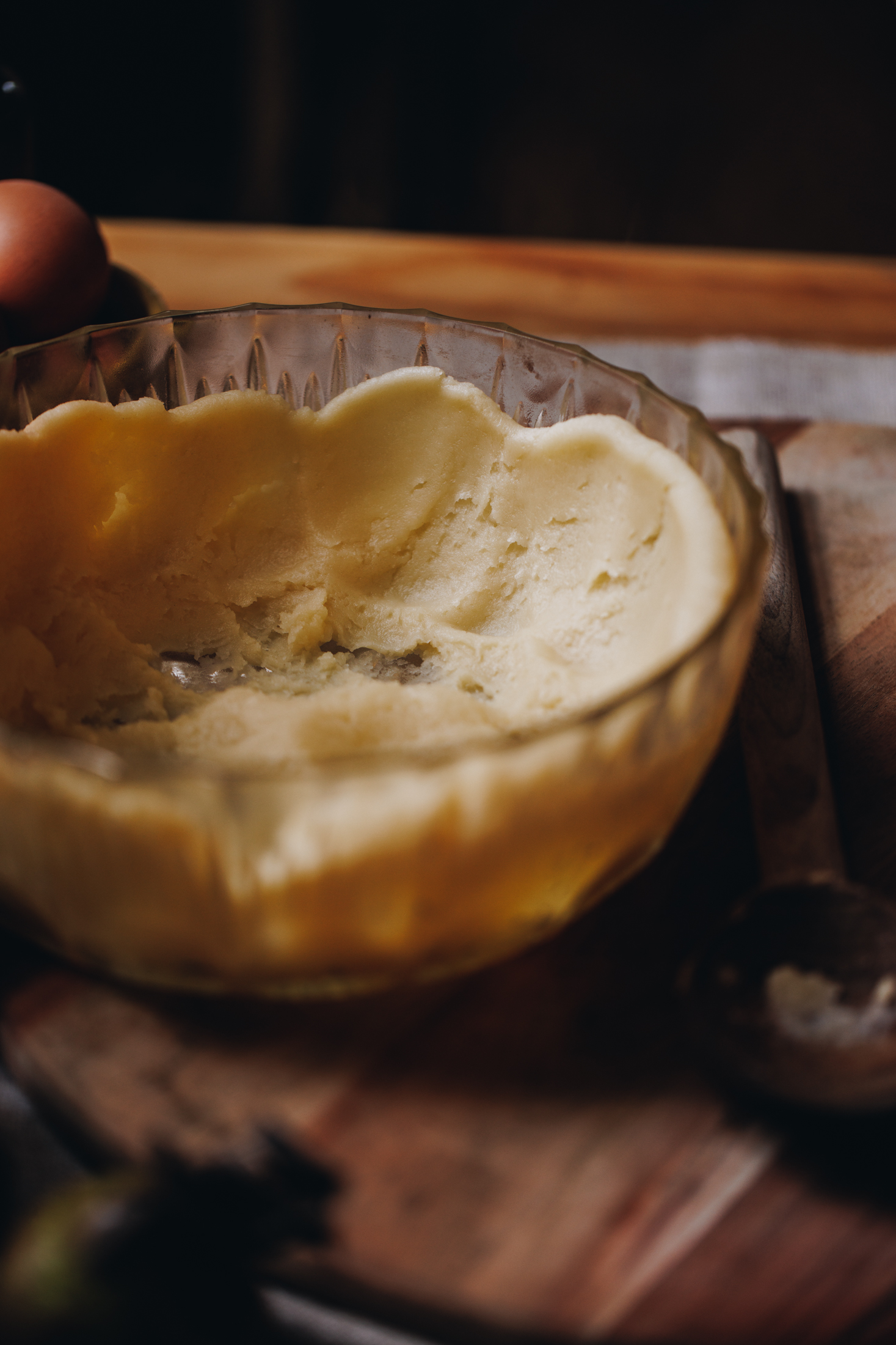 A vintage glass bowl sits on a wood board on a brown cloth on a wooden table. In the bowl is choux pastry that has been spread up the sides of the bowl so it cools down. 