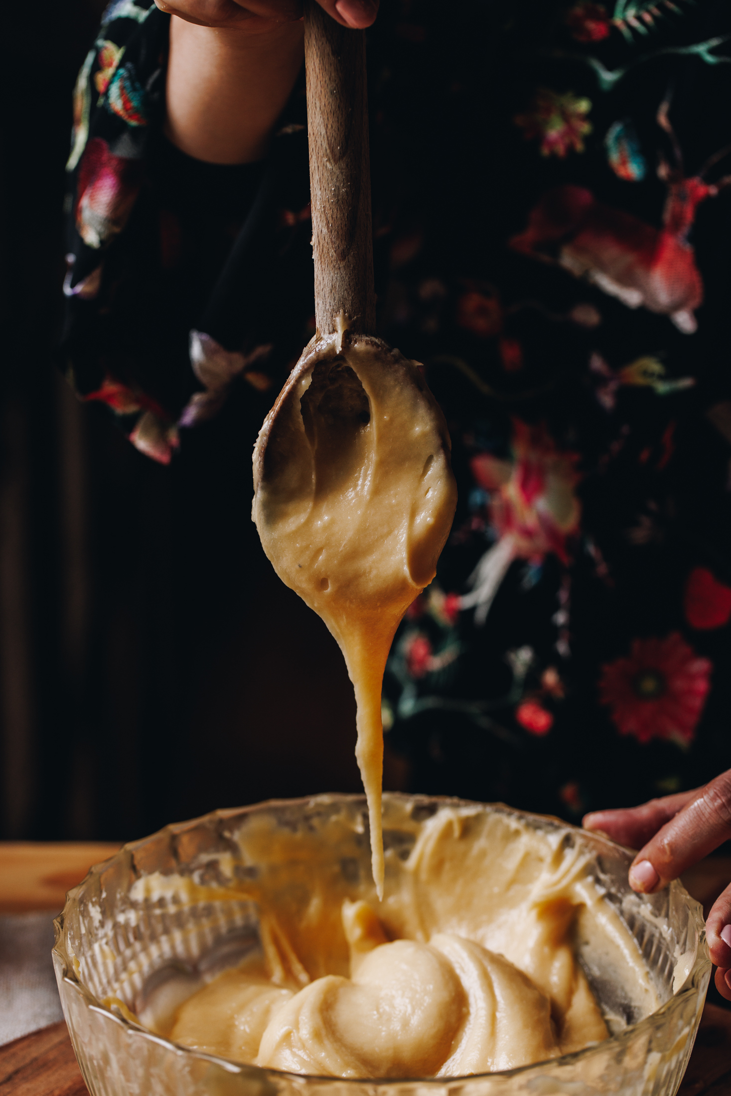 A vintage glass bowl sits on a wood board on a brown cloth on a wooden table. In the bowl is choux pastry that has been mixed with egg. A wooden is spoon has been dipped in to the mixture and a v shape is seen dripping off the end of the spoon. Naomi is holding the spoon in the background. 