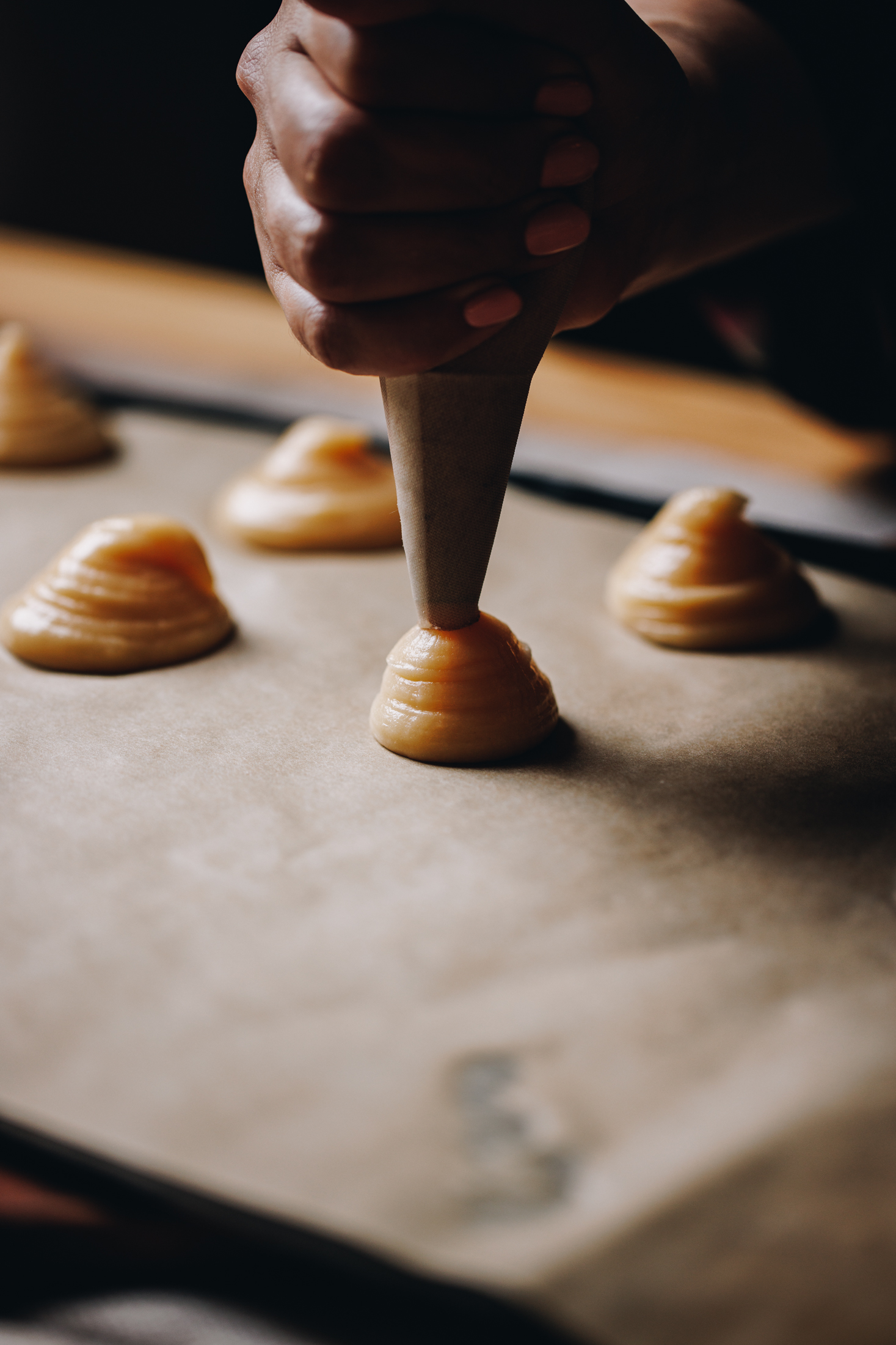 A black tray is lined with brown baking paper and is sitting on a wooden table. On the tray is blobs of choux pastry. Naomi is piping some of the pastry on to the tray. 