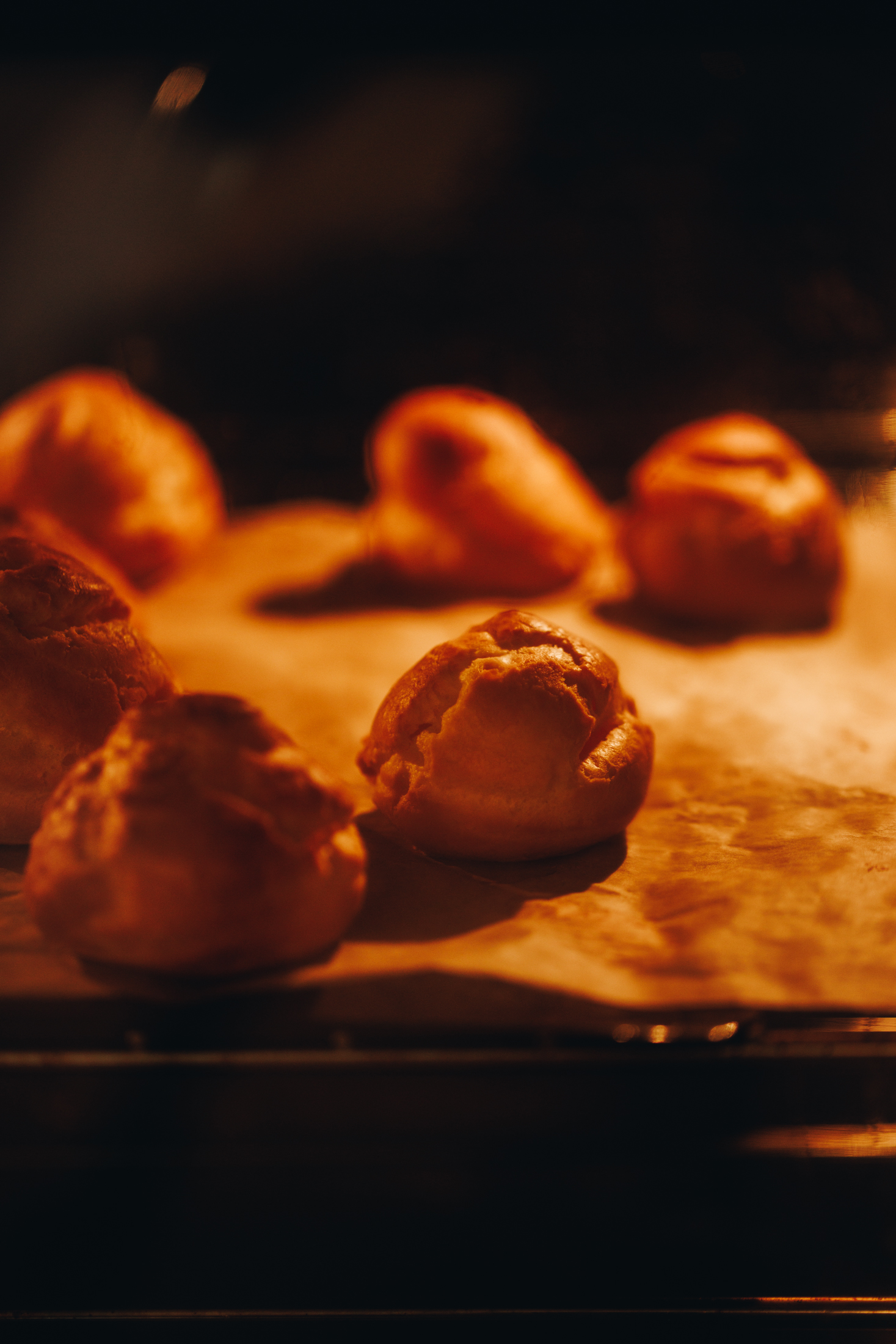 A black tray lined with brown baking paper is in the oven. Through the glass we see choux buns baking to a golden colour. 