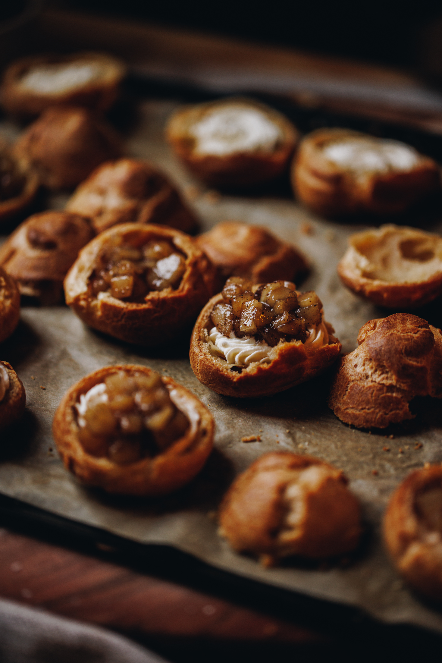 A black tray is lined with brown baking paper and is sitting on a wooden table. On the tray is baked choux pastry baked in half. Every second bun has whipped cream piped in to the base and is topped with pear compote. 