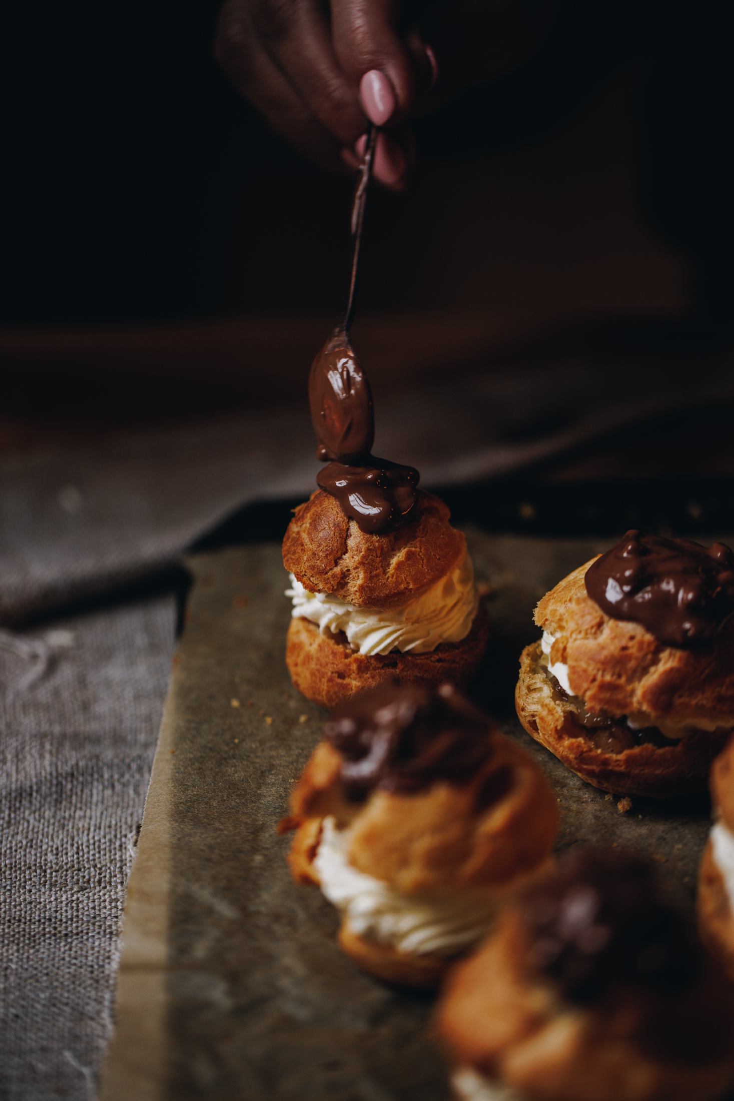A black tray is lined with brown baking paper and it sits on a grey tablecloth. On the tray is spiced pear choux buns. All of them are topped with dark chocolate and Naomi is adding the final chocolate to the last Choux bun. 