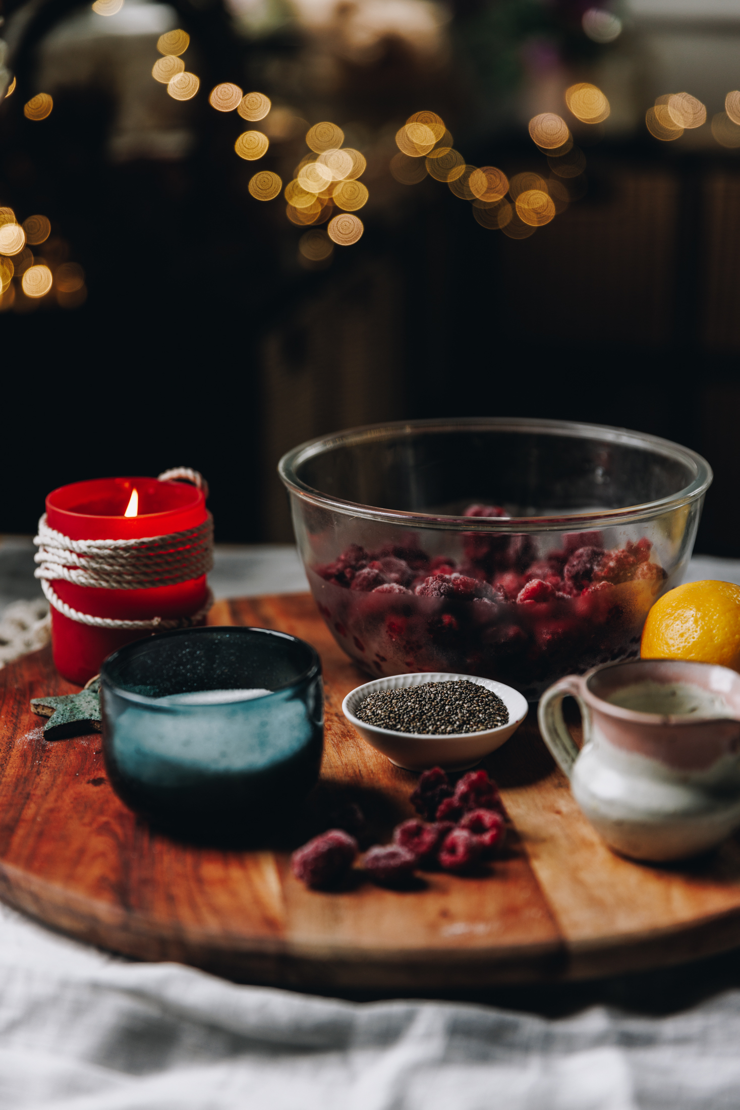 The Chia Raspberry and Lemon Jam ingredients 
are in different vintage bowls and are sitting on a wooden board. A red candle are behind the ingredients.