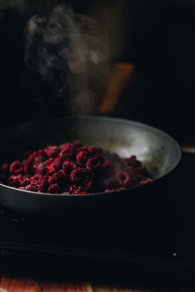 Frozen raspberries are on a frypan in a moody shot. Steam is coming off them as they are starting to heat for the Chia Raspberry and Lemon Jam.