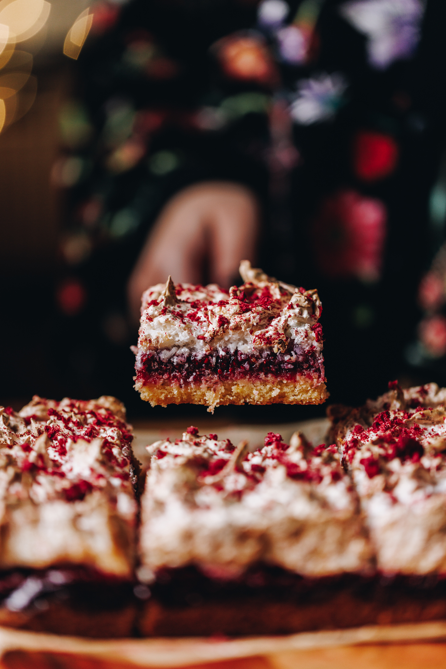 One piece of Louise Cake Slice is being held in the air to reveal the buttery base, thick raspberry slice and coconut meringue with free-dried raspberries.