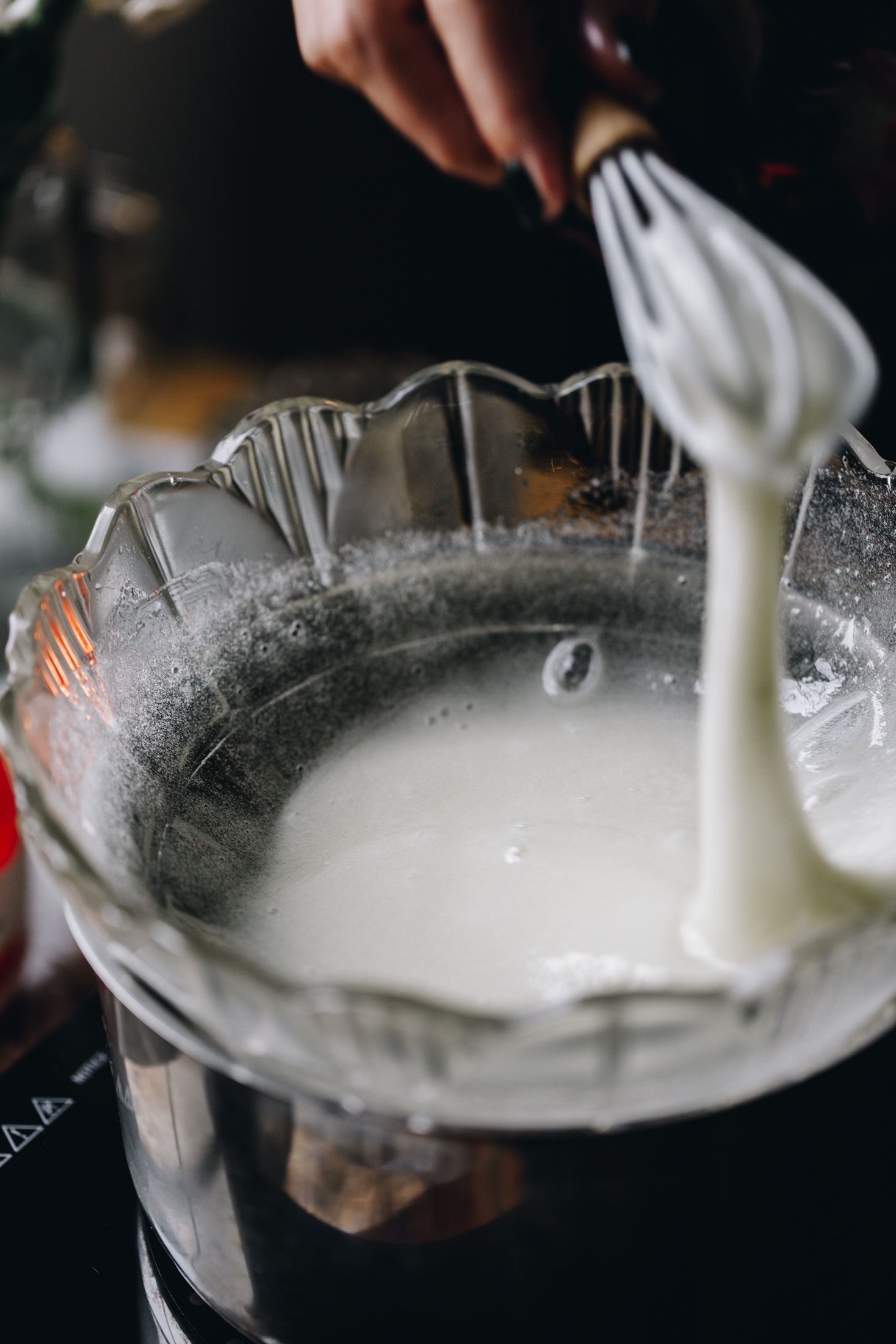 A glass bowl sits on a silver pot. Inside the bowl is egg whites that are being whisked by a small whisk, the egg whites are lifted in the air showing the consistency.