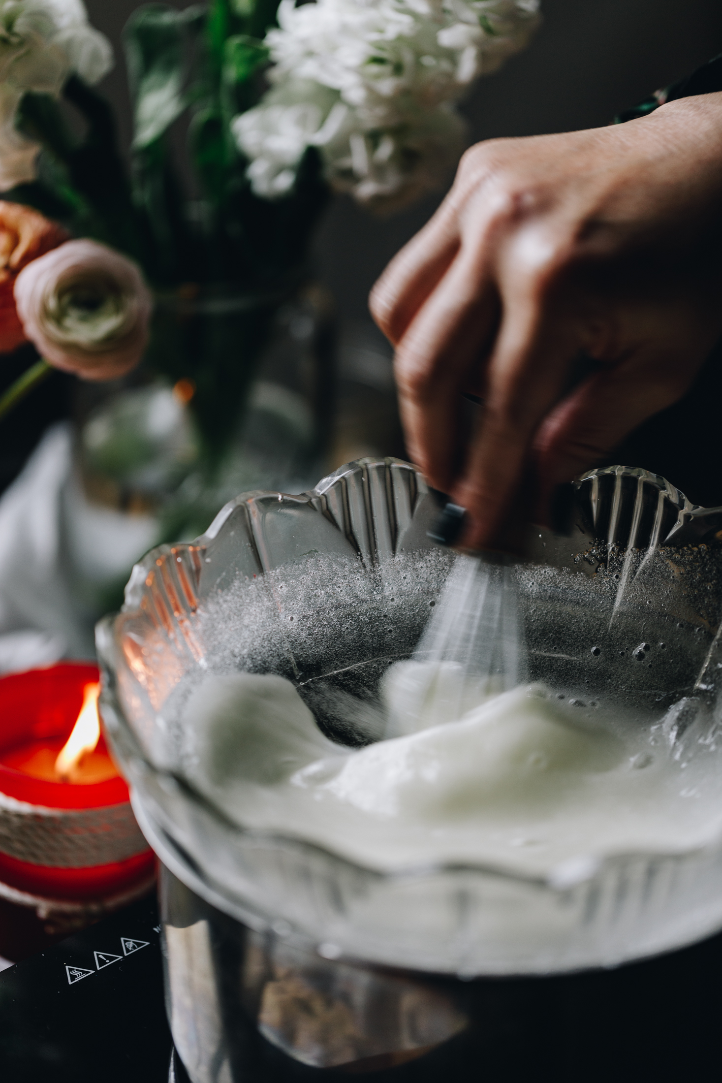 A glass bowl sits on a silver pot. Inside the bowl is egg whites that are being whisked by a small whisk. Flowers and a candle are in the background.