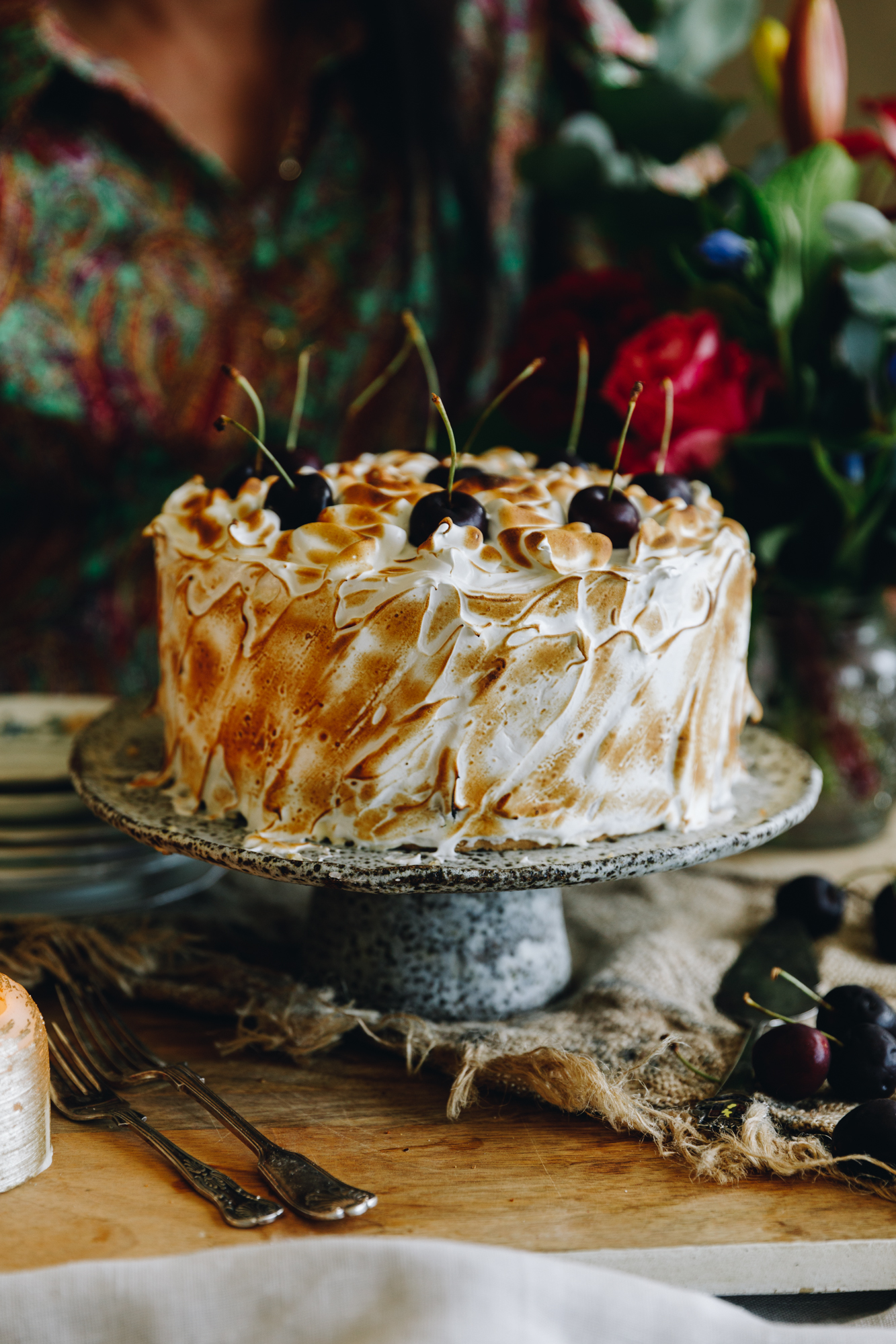 A whole Gluten-free almond sponge is on a ceramic cake stand on a wooden table. It Is covered in torched Swiss meringue and topped with whole cherries. 