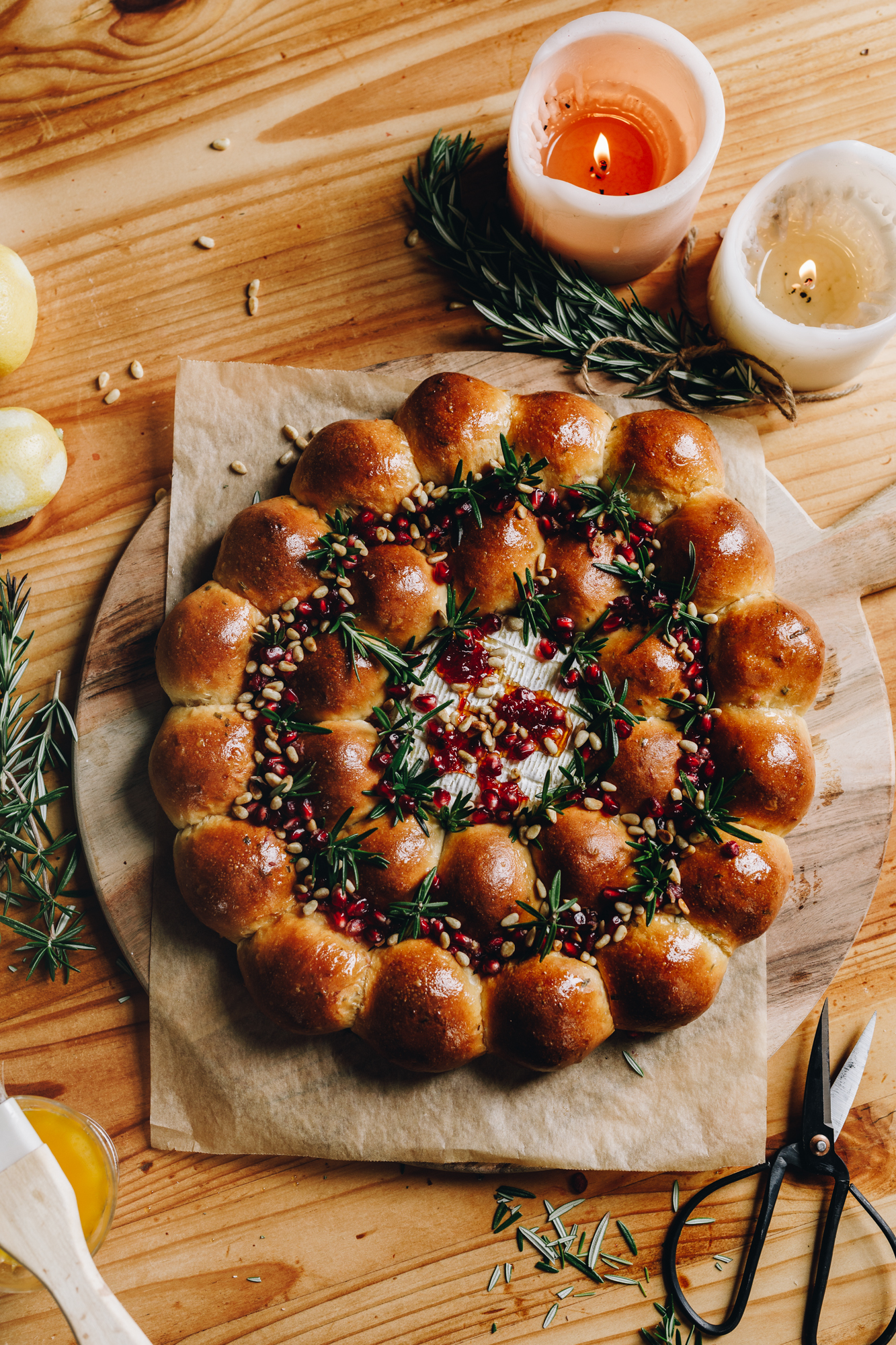 A flat lay shot reveals a freshly baked brie cheese bread wreath. It has golden baked buns that are arranged in a wreath shape that have sprigs of rosemary and pomegranate seeds scattered in between them. In the centre is baked brie cheese that has chill honey on top. The wreath is on a round wooden board, lined with baking paper. Next to it is vintage cutting scissors, two candles are burning, sprigs of rosemary and pine nuts are on the wooden table. 