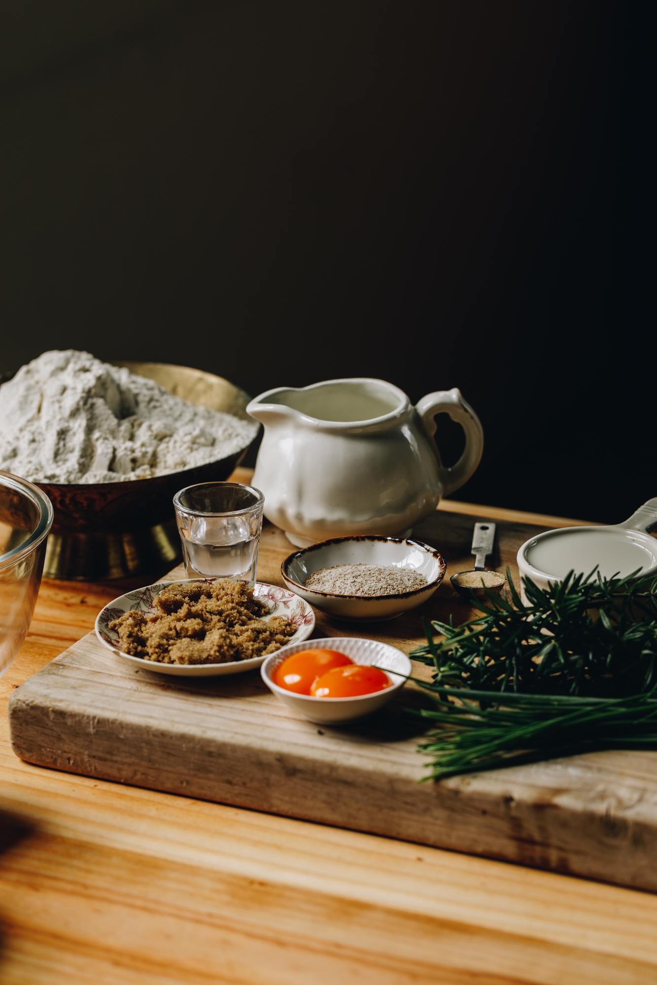 On a wooden table sits a wooden board with bread ingredients on it. In vintage cups and small bows are egg yolks, brown sugar, yeast, milk and in a brass bowl is flour. Fresh chives and rosemary are piled on the board. 