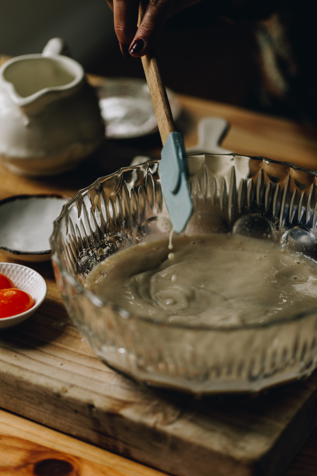 A wooden board sits on a wooden table. On the board is a glass vintage bowl that has yeast ingredients, it has been stirred with a blue spatula with a wooden handle, which is still hovering in the middle of the yeast mixture. Next to the bowl is egg yolks in a small white bowl. A white jug and a couple of white little bowls and cups are also surrounding the bowl. 