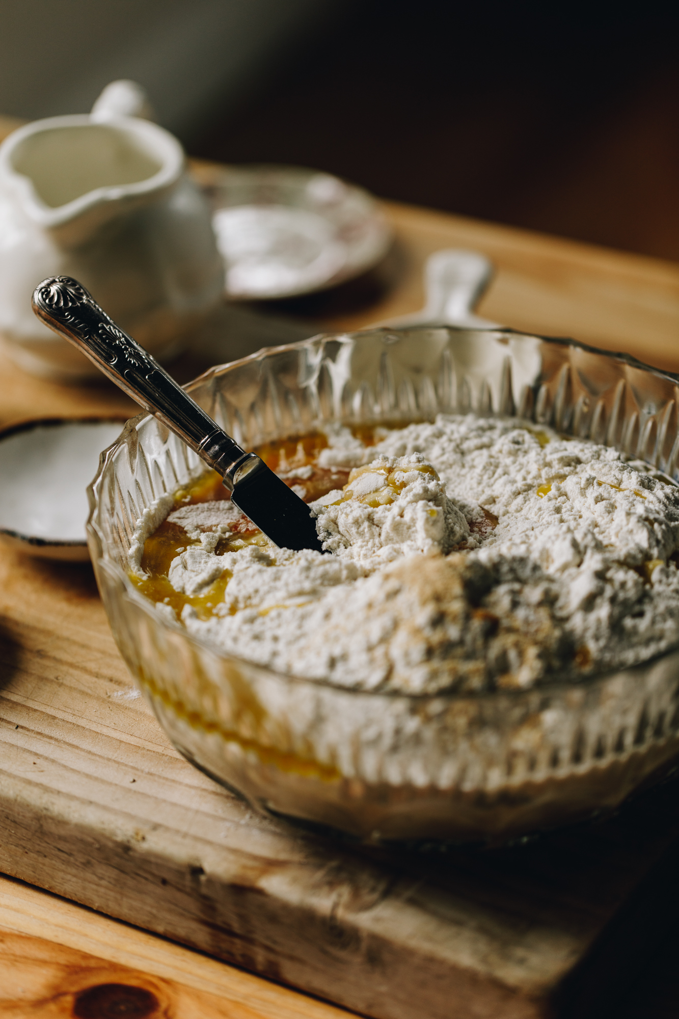 A wooden board sits on a wooden table. On the board is a glass vintage bowl that has yeast ingredients, oil and flour in it. A bread and butter knife is in the mixture, leaning on the glass bowl. Next to the bowl is a small white bowl. A white jug and a couple of white little bowls and cups are also surrounding the bowl.