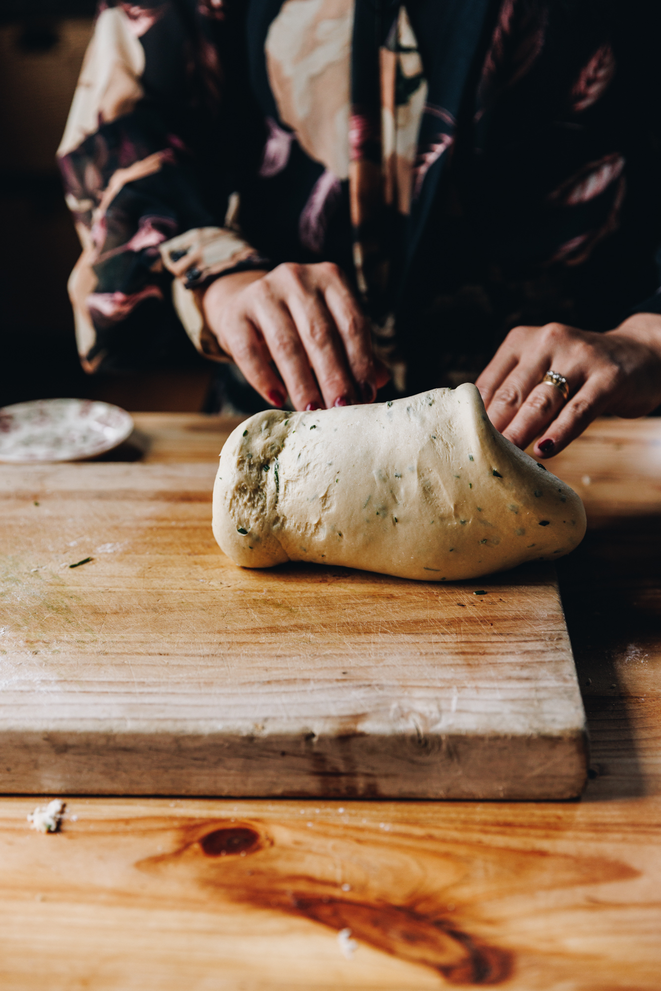 A wooden board sits on a wooden table. On the board Naomi is kneading the bread dough. The dough is speckled with green herbs. A little vintage plate is in the background. 