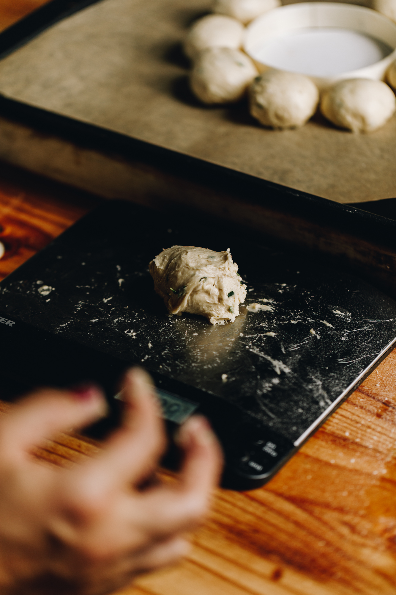 On a wooden table sits a silver scale that is weighing a piece of bread dough. In the background is a black tray, lined with baking paper with small buns arranged around a circle. 