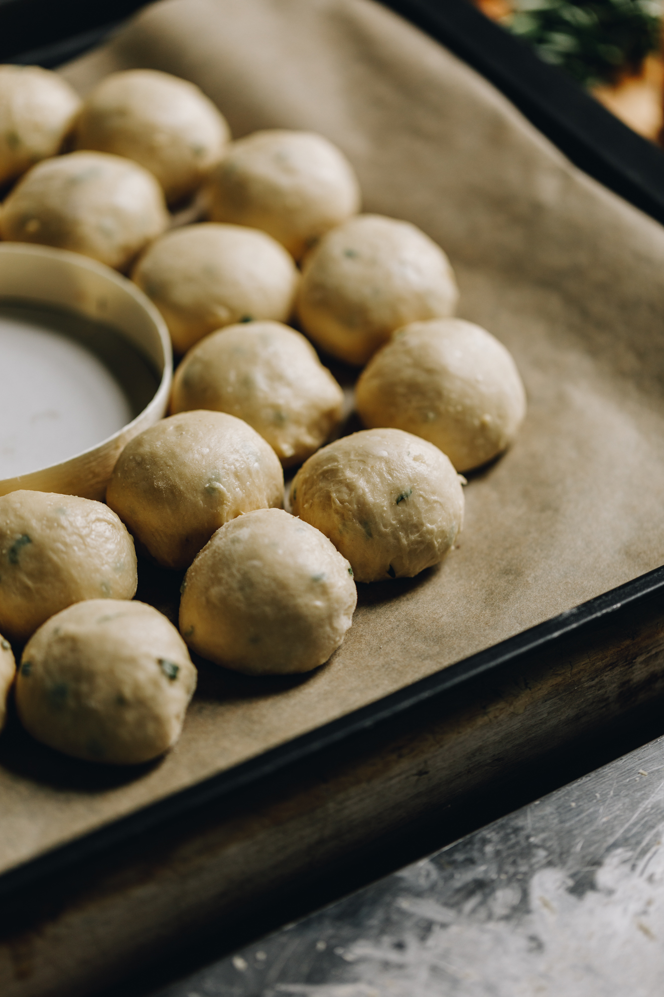 On a black tray, lined with brown baking paper is round Brie cheese packaging in the centre. Surrounding the packaging is small herbed bread buns that are shaped in a wreath.