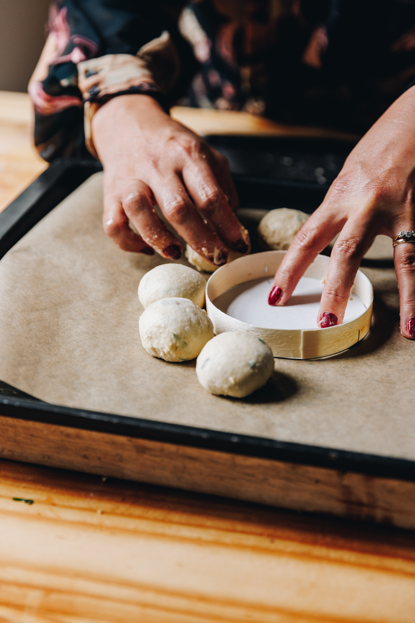 On a black tray, lined with brown baking paper Naomi is placing a round Brie cheese packaging in the centre. Surrounding the packaging is three, small herbed bread buns