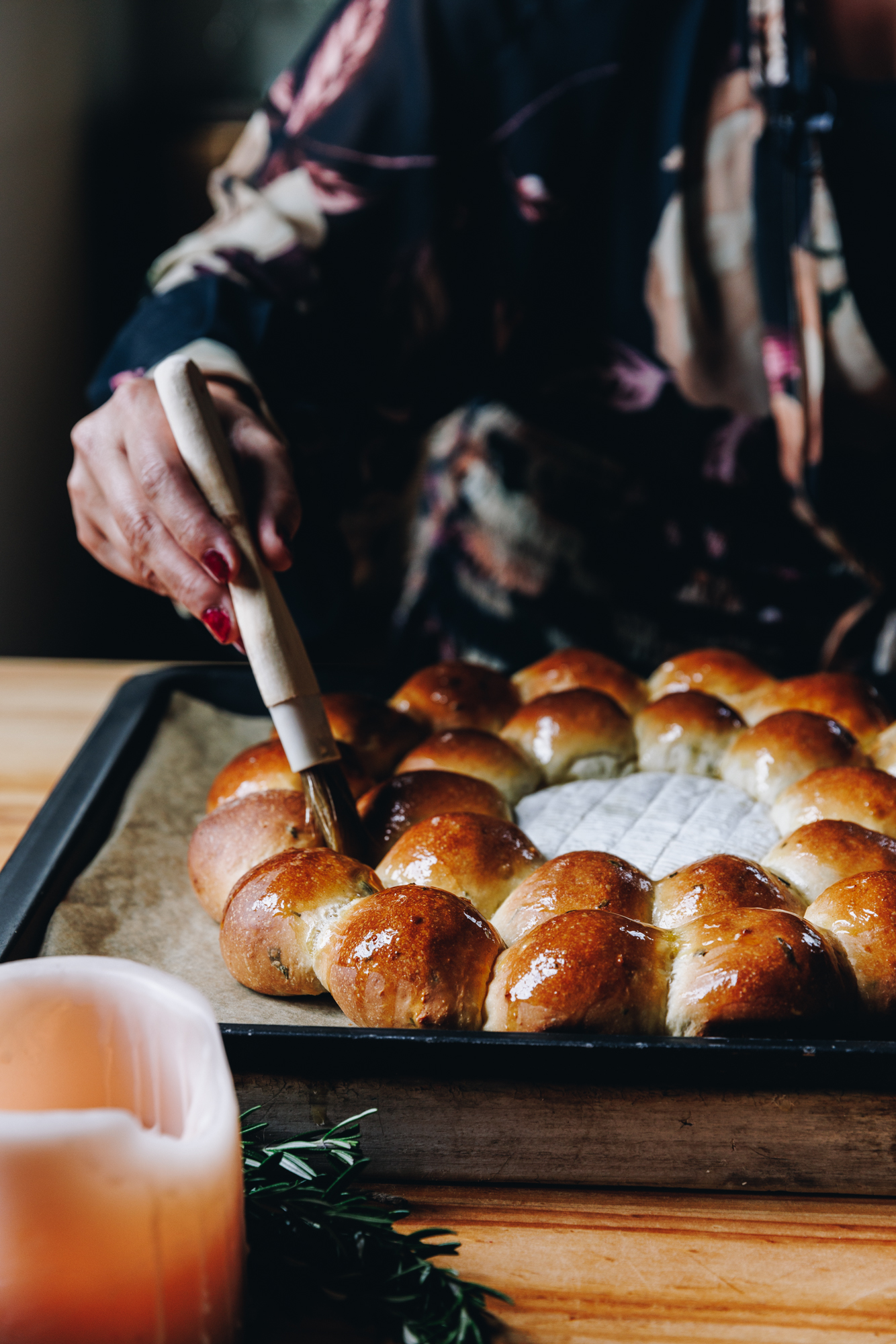 On a black tray, lined with brown baking paper is a freshly baked Brie cheese bread wreath. It is being glazed with melted butter by Naomi, she is using a pastry brush. In the forefront of the shot is a burning and a a couple of sprigs of fresh rosemary. 