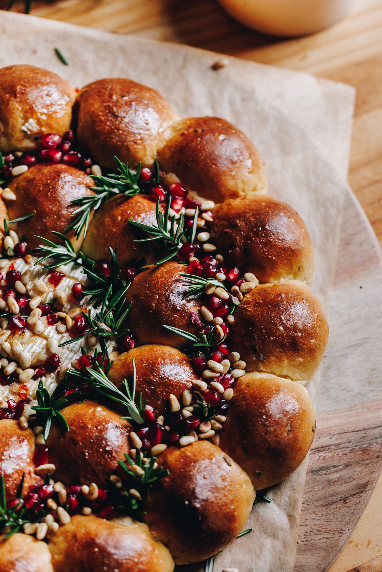A round wooden board lined with brown baking paper sits on a wooden table. On the board is a freshly baked brie cheese bread wreath that has been glazed with butter.  The bunds are arranged in a wreath shape that have sprigs of rosemary and pomegranate seeds scattered in between them. In the centre is baked brie cheese that has chill honey on top. 