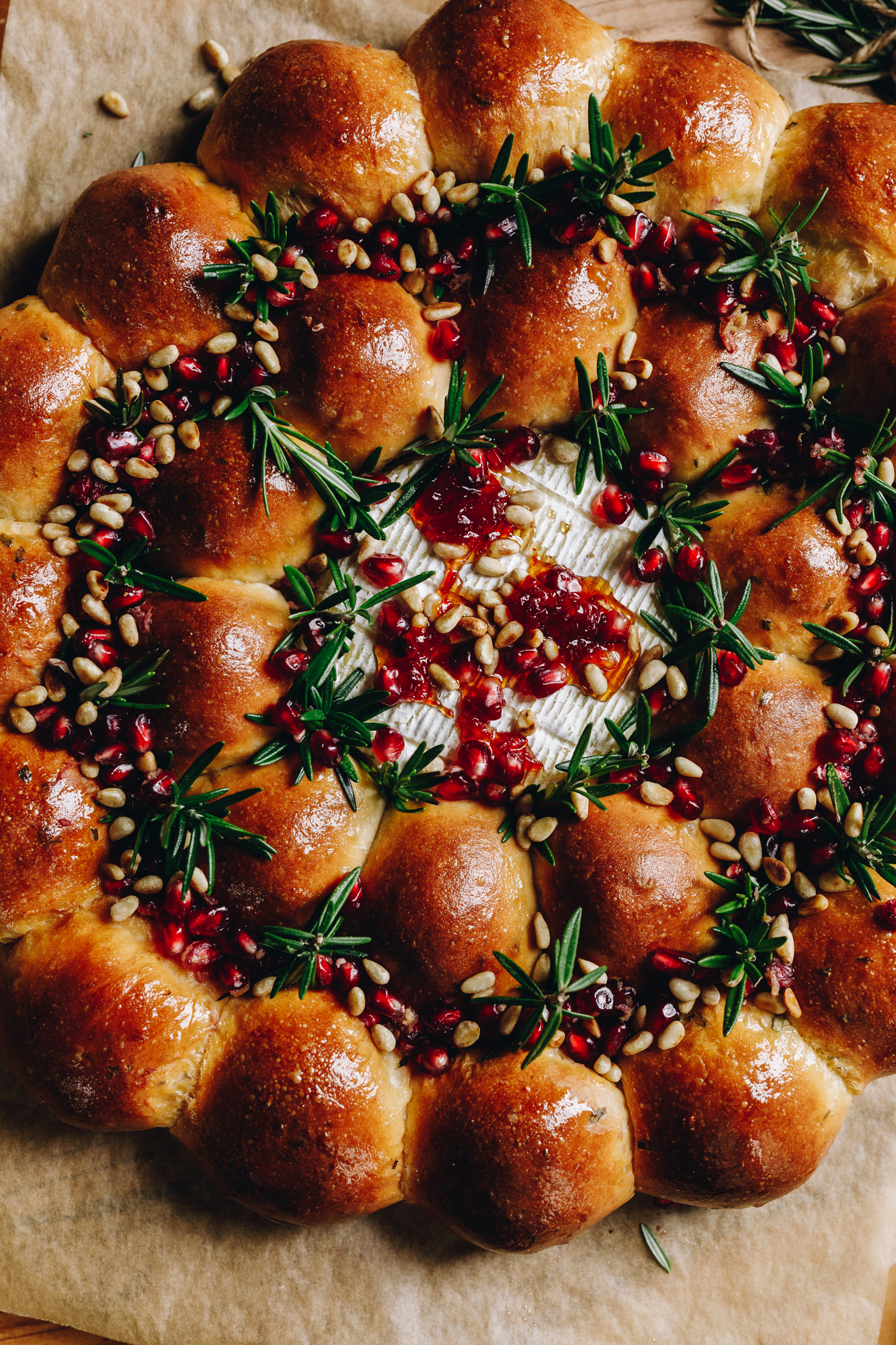 A round wooden board lined with brown baking paper sits on a wooden table. On the board is a freshly baked brie cheese bread wreath that has been glazed with butter. The bunds are arranged in a wreath shape that have sprigs of rosemary and pomegranate seeds scattered in between them. In the centre is baked brie cheese that has chill honey on top.