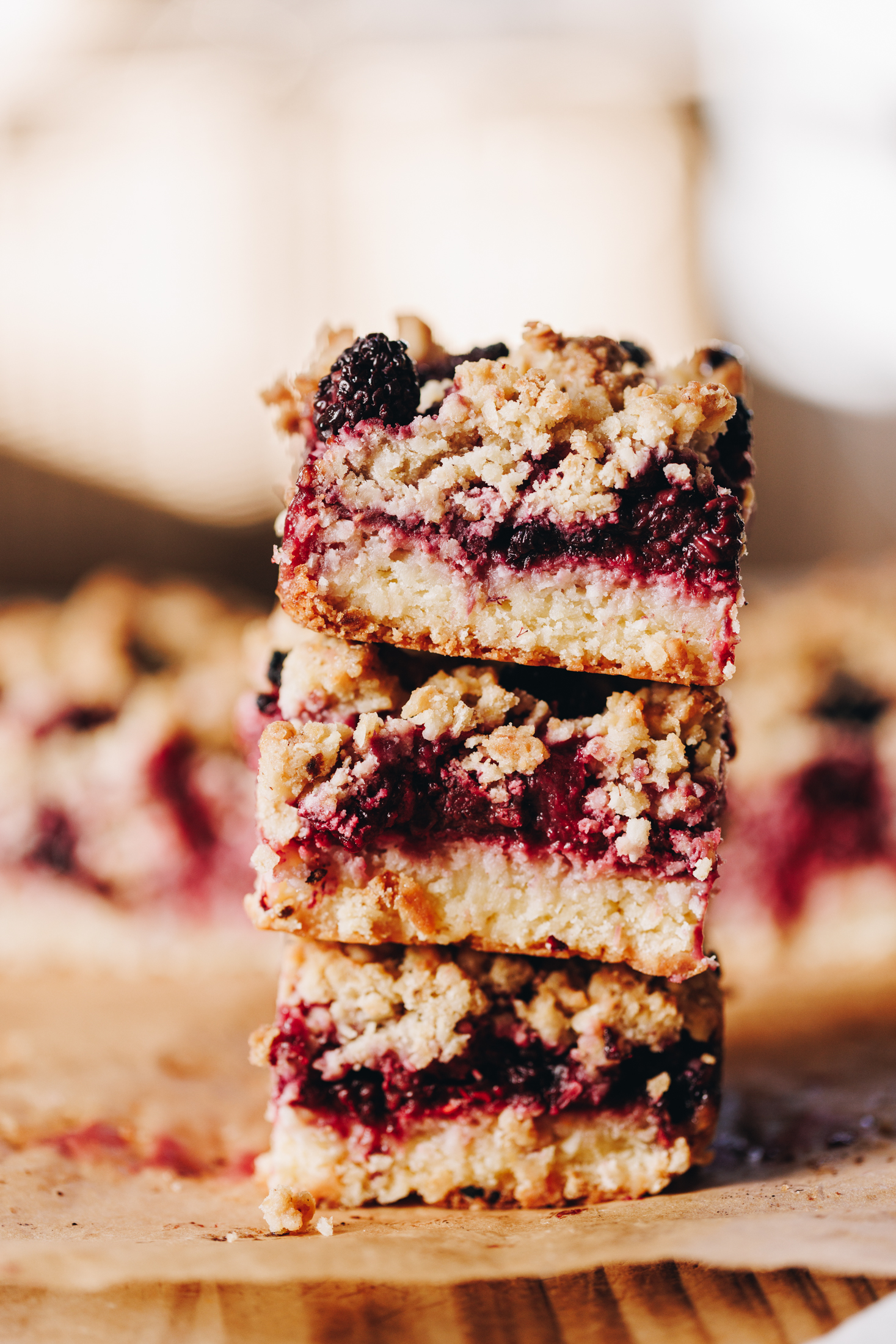 A wooden board is lined with brown baking paper. Three freshly baked squares of berry oat crumble slice have been stacked on top of each other and show the buttery base, the berry centre and the crumble topping of the slice. 