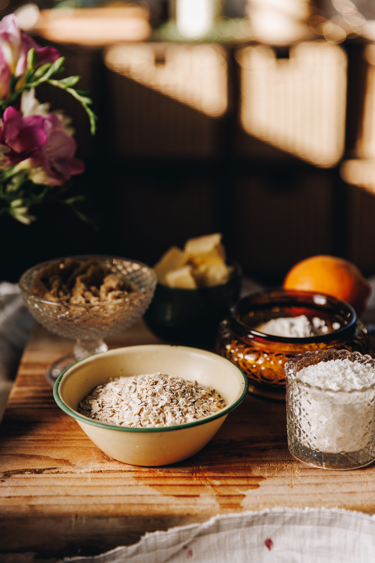 On a wooden table is a wooden board that is sitting on a beige cloth. Pink flowers are in a glass jar next to the board. On the board is vintage bowls and cups. There is oats, coconut, sugar, brown sugar, butter and an orange in view.
