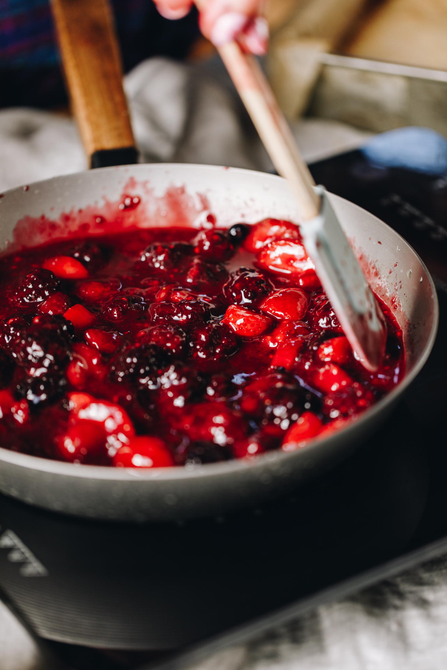 A grey frying pan sits on a black portable stove. In the pan is mixed berries the are cooking in a sauce and are being stirred with a blue spatula with a wooden handle. 