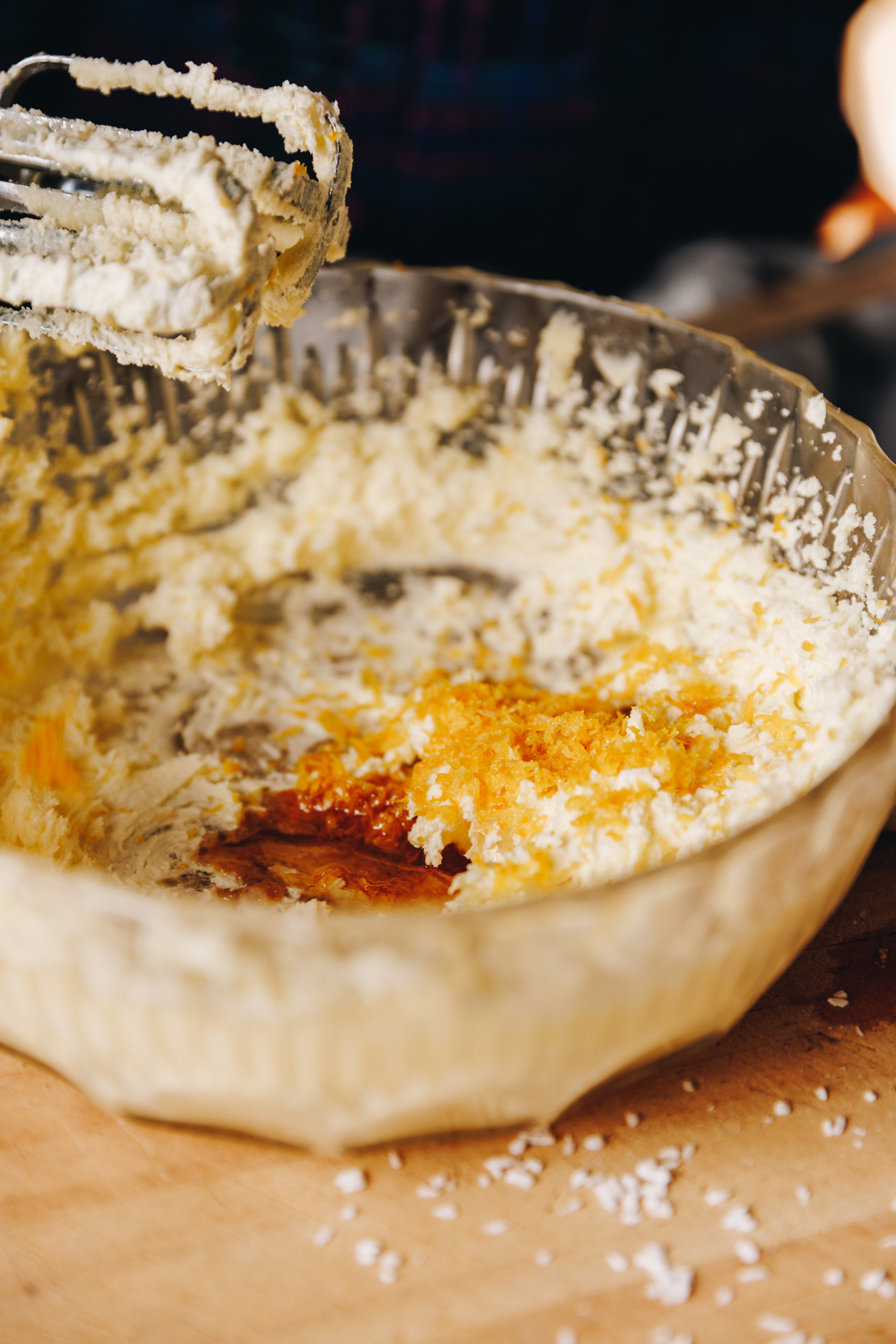 A glass vintage bowl sits on a wooden board. In the bowl is whipped butter that has had finely grated lemon zest and vanilla added to it. Next to the bowl is a stand mixer whisk attachments. 