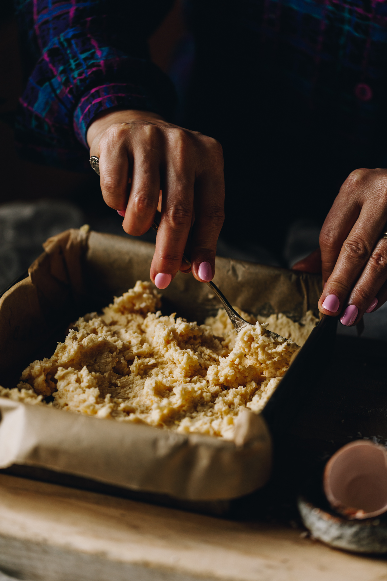 A vintage baking tin lined with brown baking paper is on a wooden board. In the tin is a coconut and butter mixture that Naom is spreading with a spoon. 