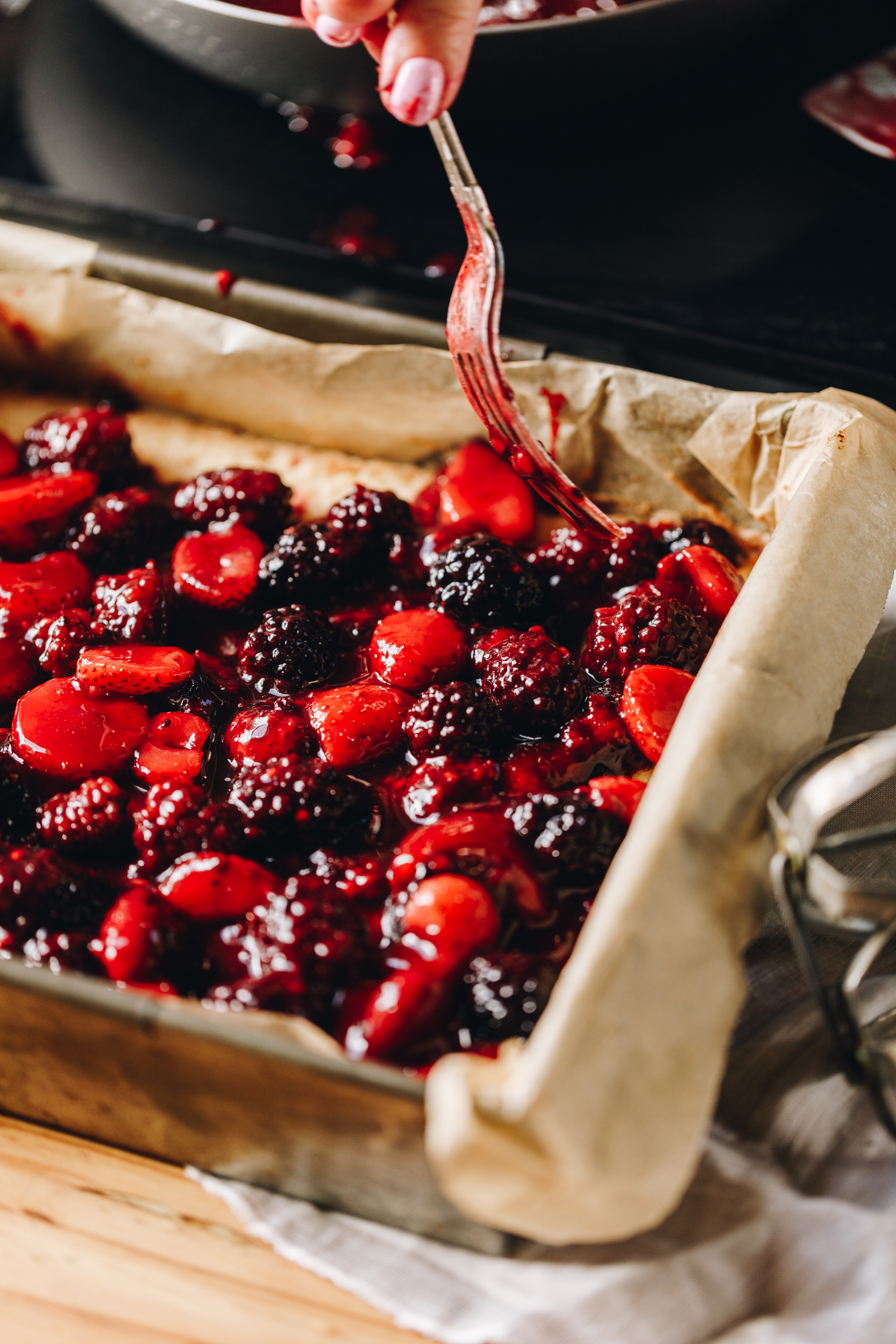 A vintage baking tin lined with brown baking paper is on a wooden board. In the tin is a biscuit base that has been covered with mixed berries. A silver vintage fork is arranging them in to place.
