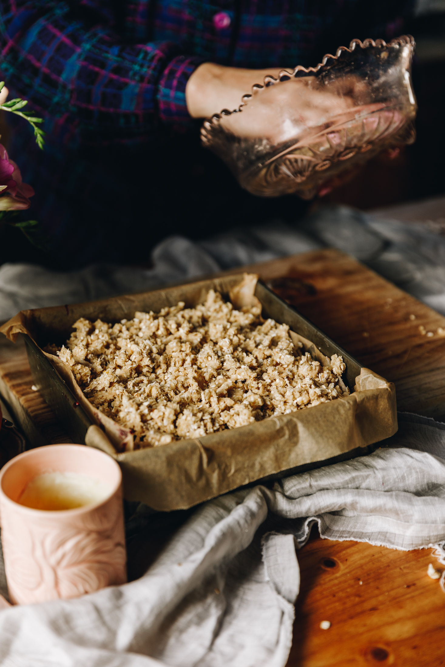 A vintage baking tin lined with brown baking paper has a berry oat crumble slice in it. Naomi has her hand in a vintage bowl behind it, gathering the last bits of oat crumble. A soft grey tablecloth is under the tin and a peace candle is next to the tin along with a peep of flowers coming through. 