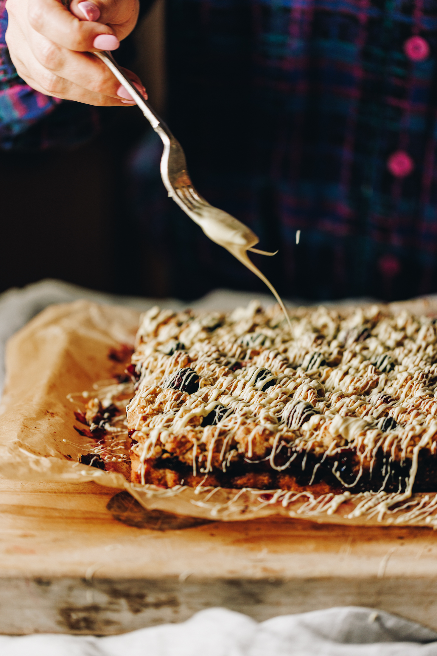 A baked berry oat crumble slice is on a wooden board that is lined with brown baking paper. Naomi is using a vintage fork to drizzle melted white chocolate on top of it. 
