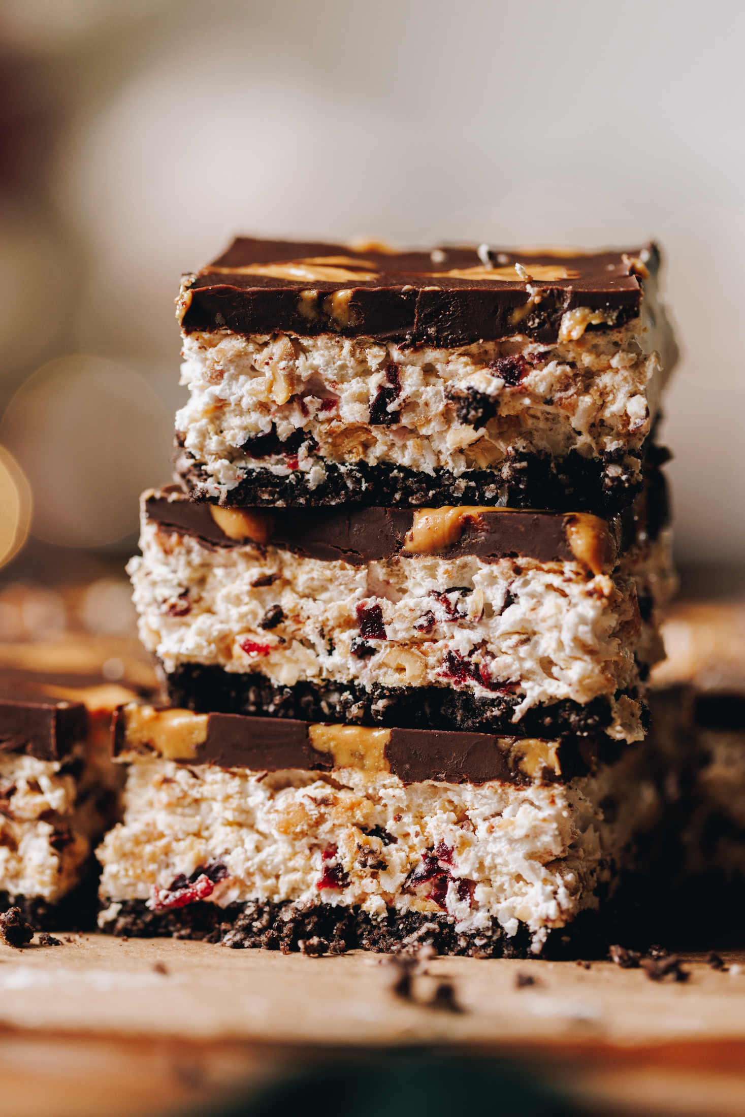 Three pieces of no-bake marshmallow slice are stacked on top of each and sit on a wooden board. The slices show the biscuit base, the textural marshmallow centre and the chocolate topping with a peanut butter drizzle.
