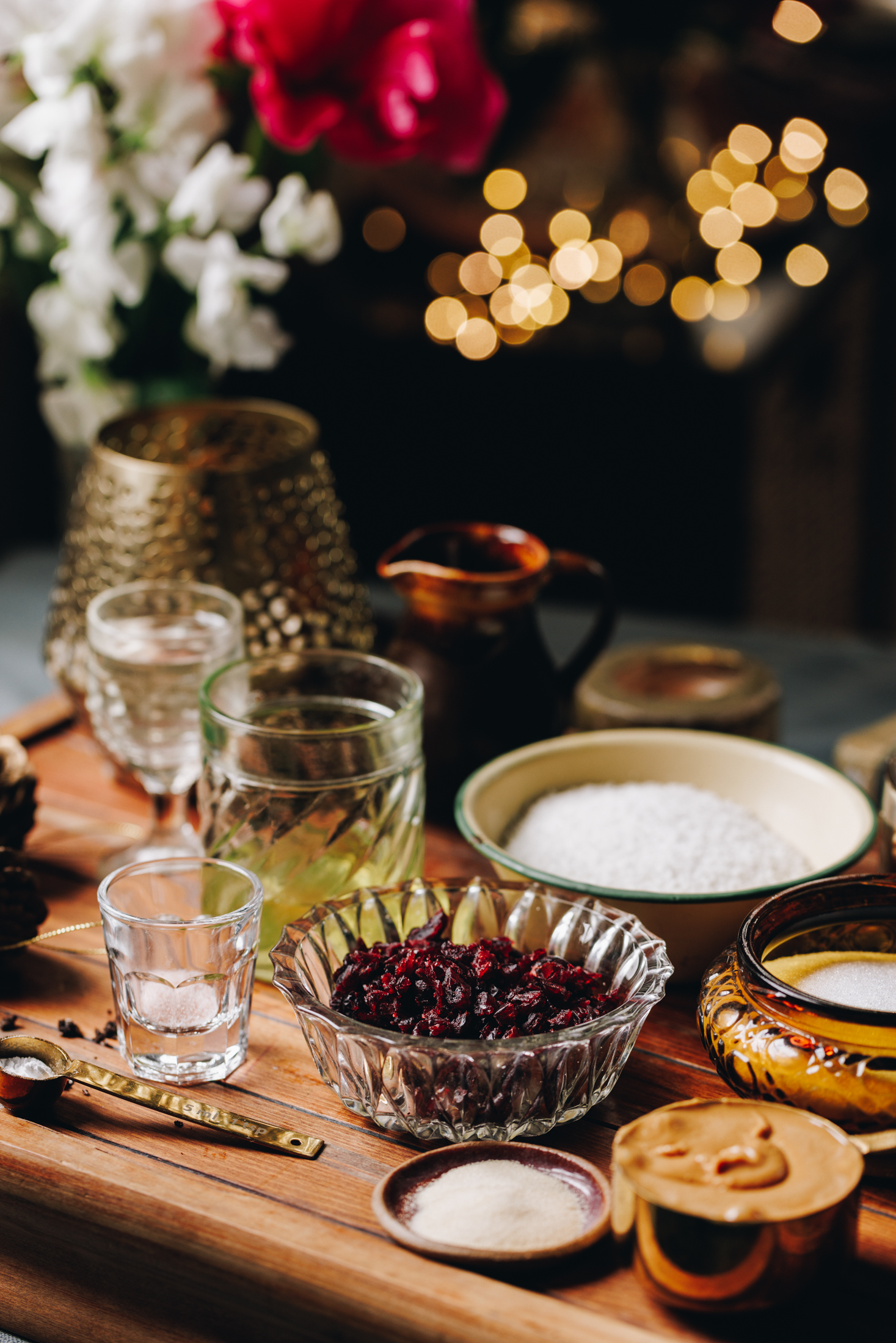 A wooden board sits on a table. It is filled with vintage bowls, spoons and jars that have slice ingredients in them. There is egg whites, caster sugar and cranberries in view. A vase of flowers is in the background with golden fairy lights. 