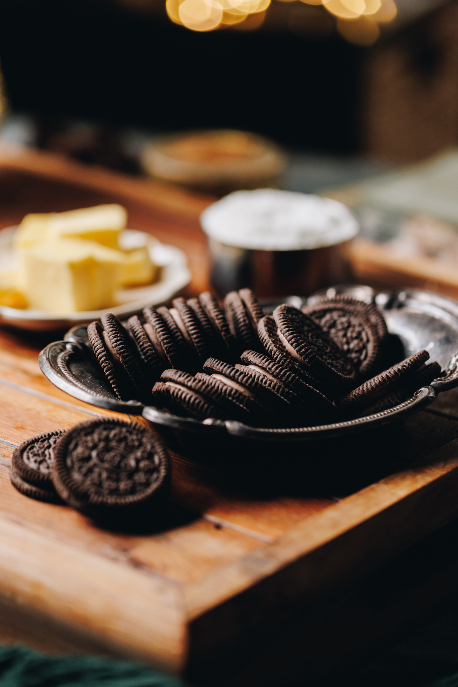 A wooden board sits on a table, on the board is a vintage silver bowl that has chocolate Oreo cookies in it. Behind that is a silver cup of coconut and a small plate of chopped butter. Golden fairy lights glow in the background. 