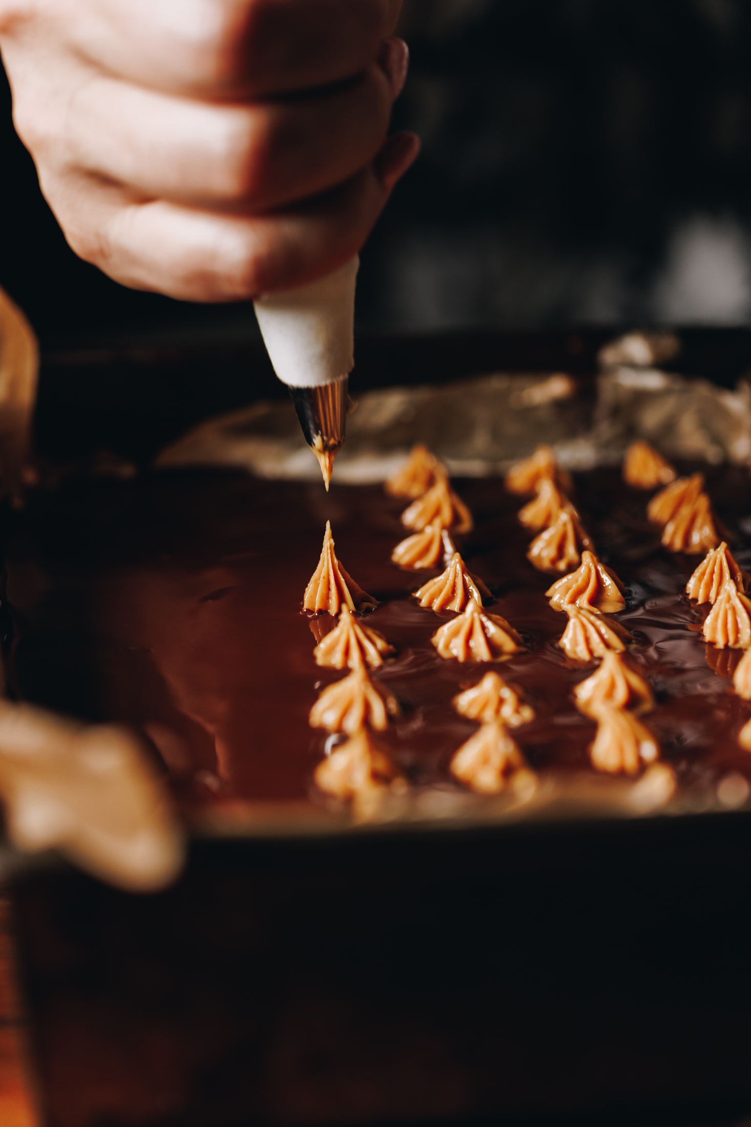A close up shows the no-bake marshmallow slice with dark chocolate on top that is still wet. On top is rows of dots of peanut butter that have been piped with a star tip. Naomi has just finished piping a dot and the peak of the peanut butter is seen. 