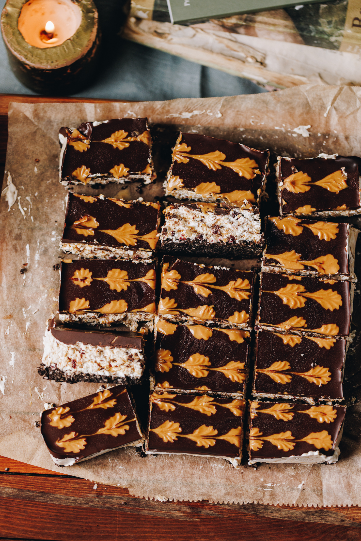A flat lay shows a no-bake marshmallow slice that has been cut in to pieces to reveal the chocolate topping with the peanut butter design. Two slices has been flipped over to reveal the layers of biscuit base, marshmallow and chocolate topping. A candle burns in the corner. 