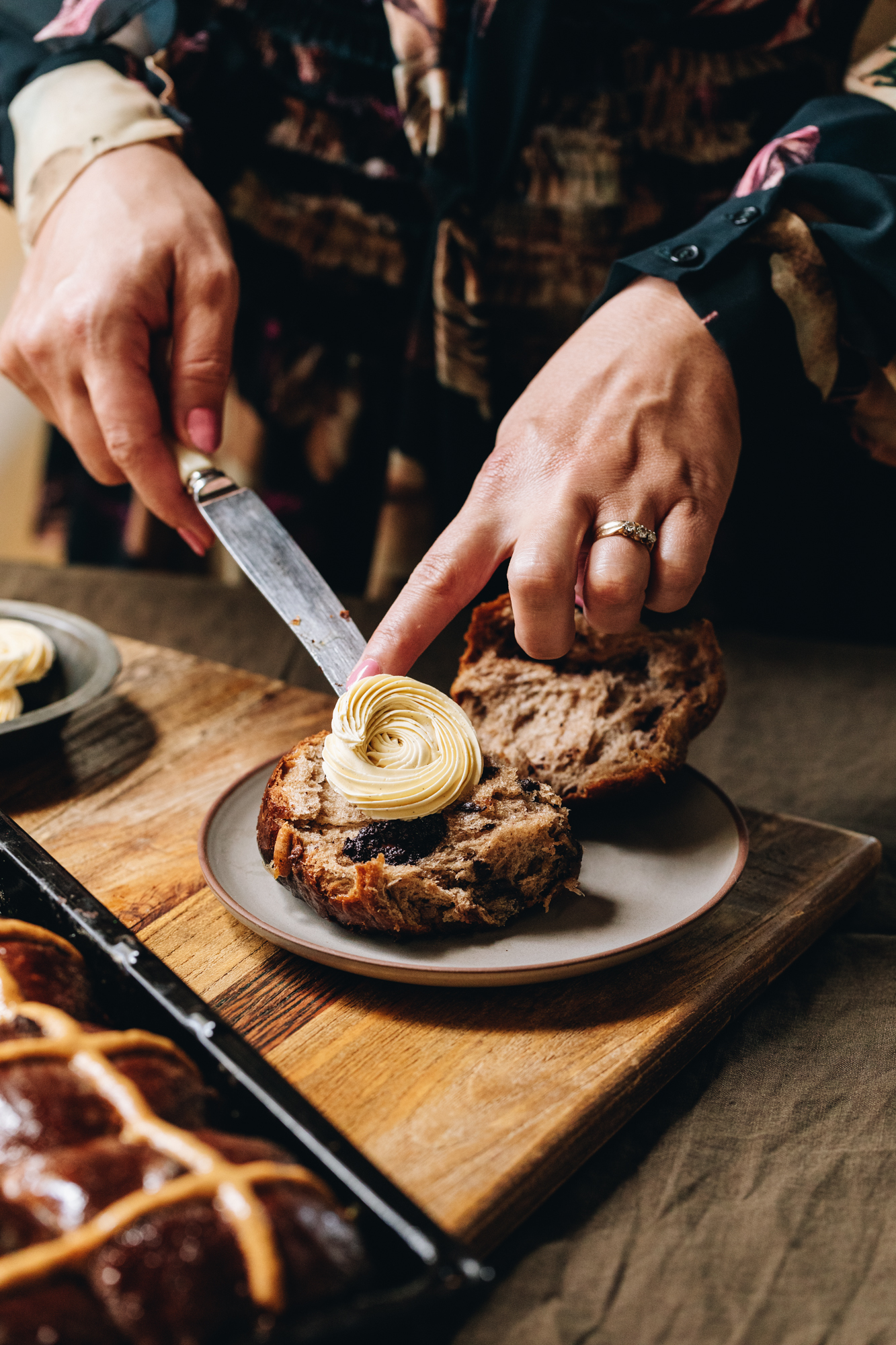 A wooden board sits on a wooden table. A brown ceramic plate is on the board and a hot cross buns is cut open. Naomi is using a knife to place the piped whipped vanilla butter recipe on to the hot cross buns. A tray of hot cross buns sits on the board next to the plate. 